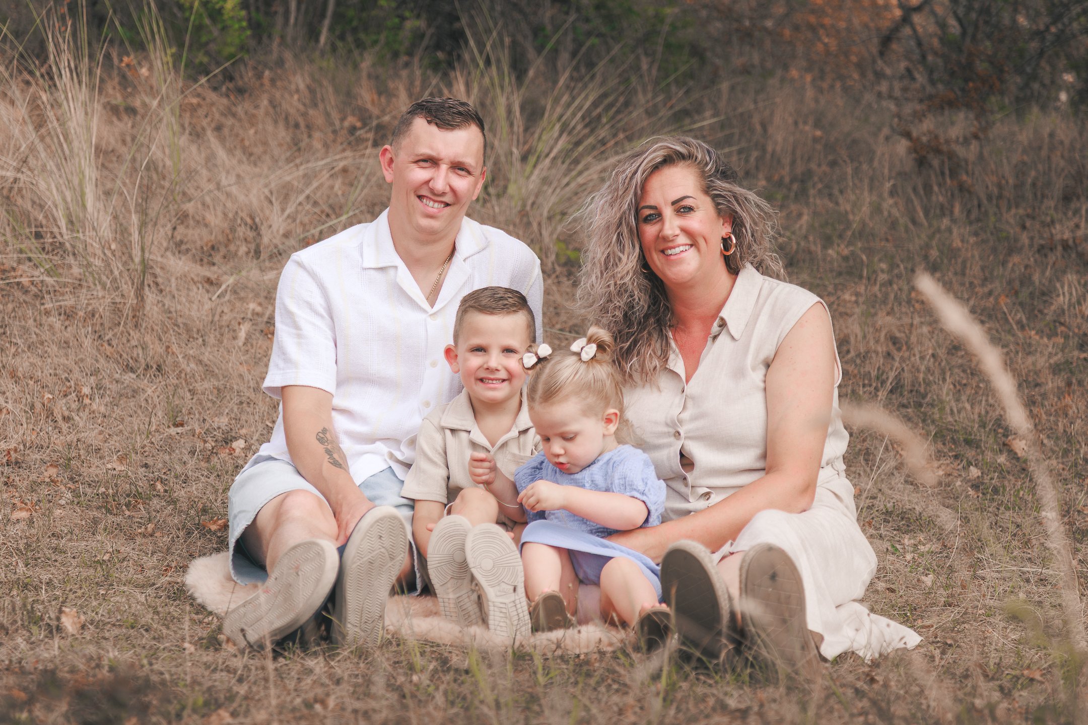 Een gelukkige familie met twee ouders en drie jonge kinderen zit samen op de grond in een natuurlijk landschap met droog gras en struiken.