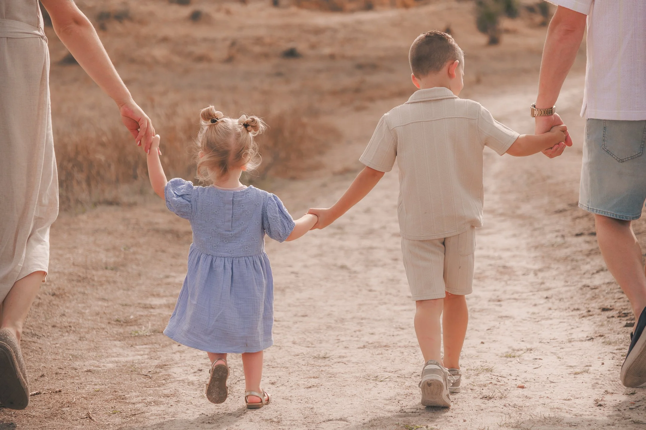 Twee jonge kinderen, een jongen en een meisje, houden elkaars handen vast terwijl ze samen wandelen op een stoffig pad in een landschap met droge begroeiing. Ze worden begeleid door twee volwassenen, waarvan alleen de armen en handen zichtbaar zijn.