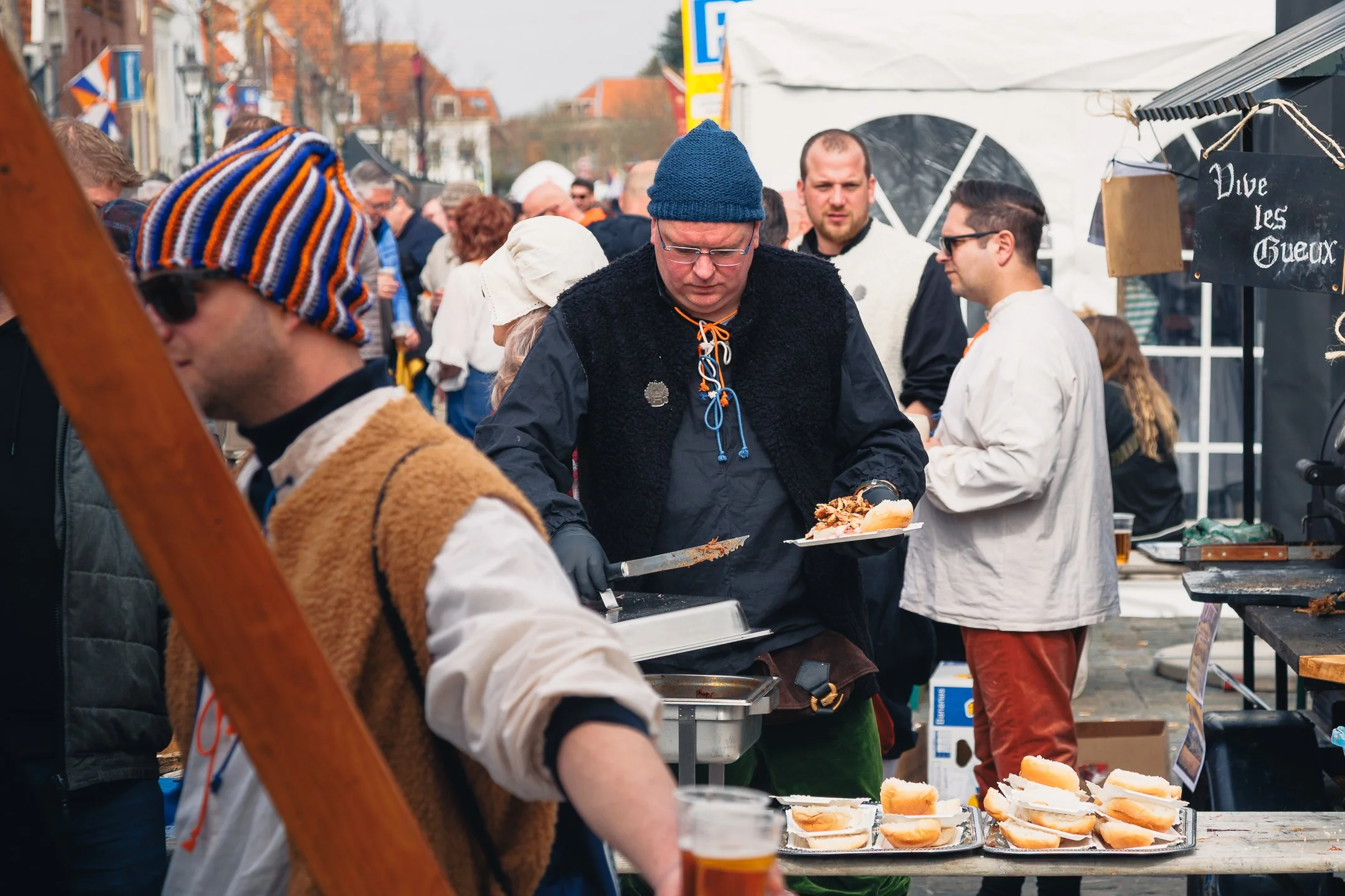 Mensen genieten buiten van een straatmarkt, waarbij ze hotdogs en dranken eten. Er is een menubord met de tekst 'Vive les Gueux'.
