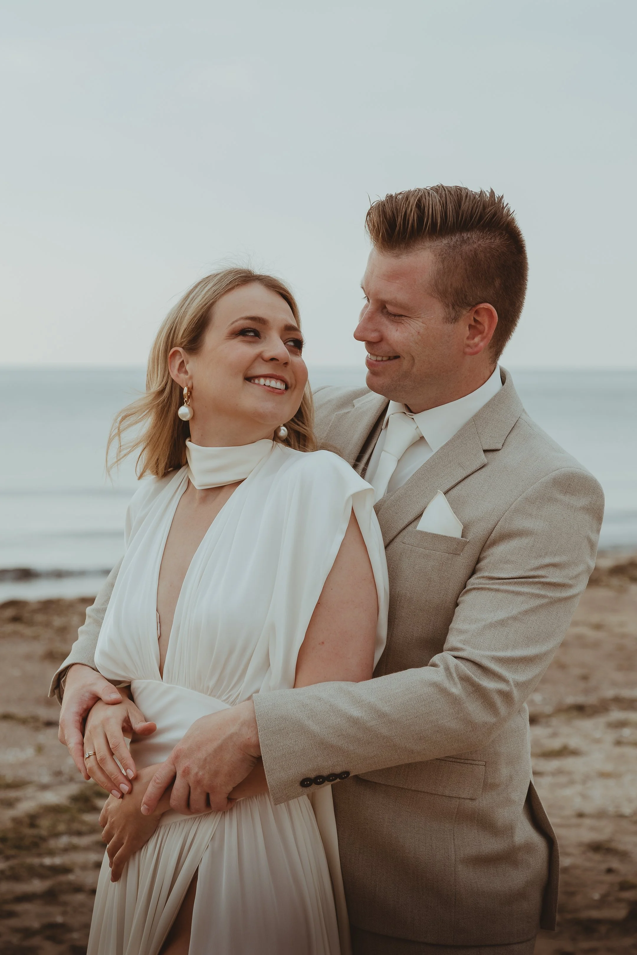 Een stel in bruiloftskleding die dicht bij elkaar op het strand staan, lachend en in een liefdevolle omhelzing, met de zee op de achtergrond.