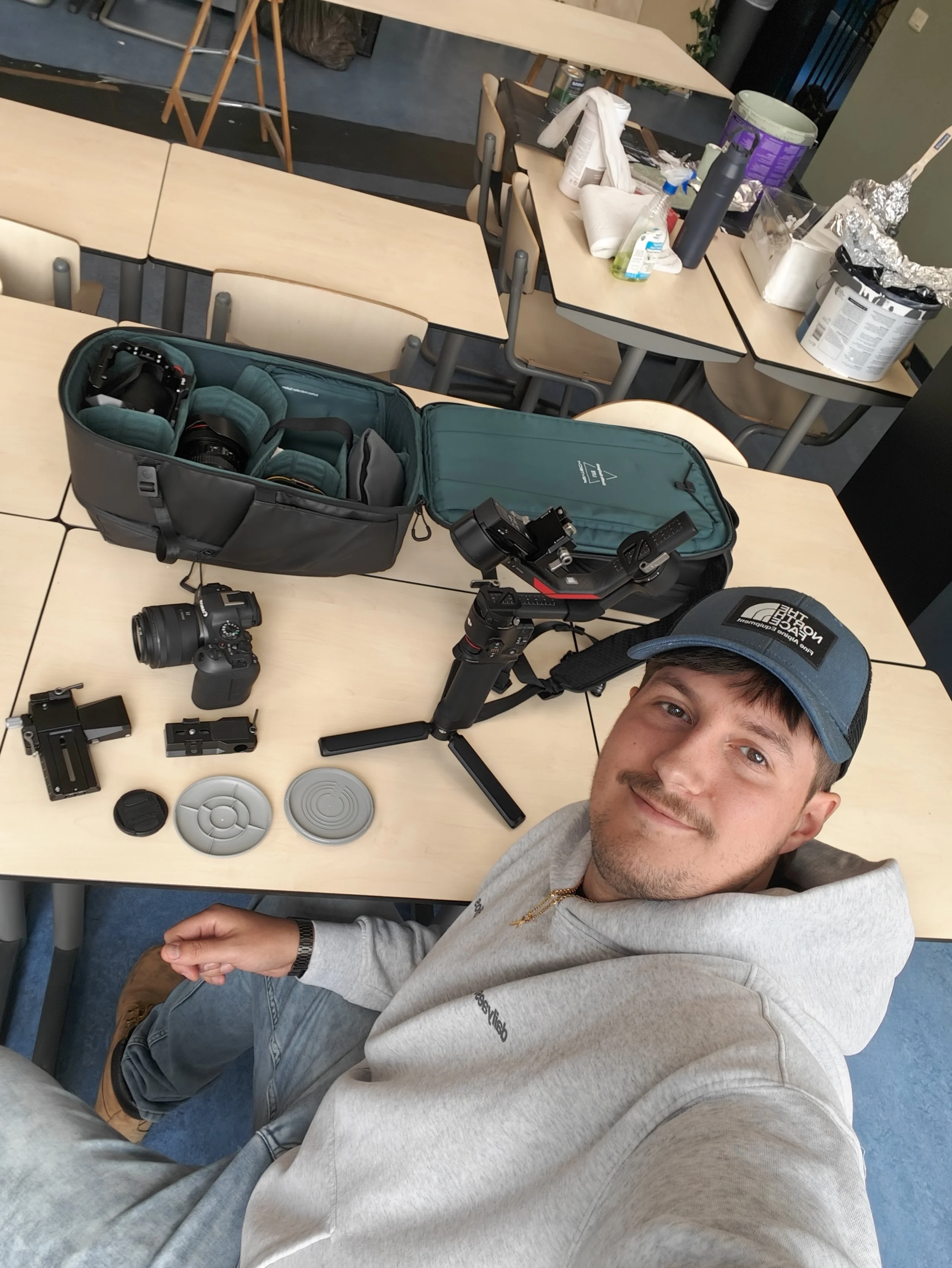 Een jonge man met een pet en een grijze hoodie liggend op een schoolbank, met fotospullen en accessoires, in een klaslokaal.