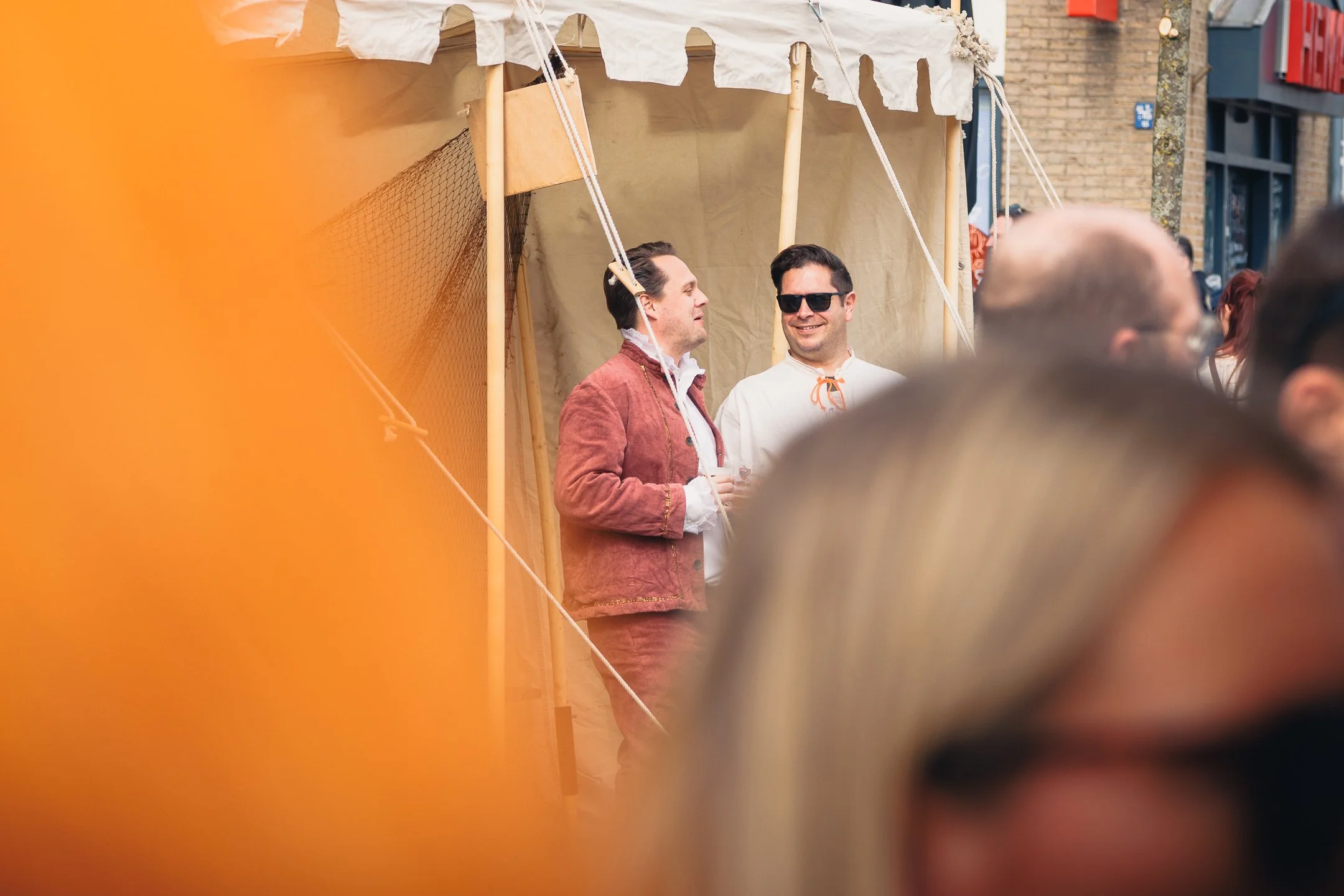 Twee mannen staan tegenover elkaar, één draagt een bruin jack en de ander een witte hoodie met een zonnenbril, onder een wit katoenen tent in de straat met een bakstenen gebouw op de achtergrond. Vermoedelijk op een markt of festival.