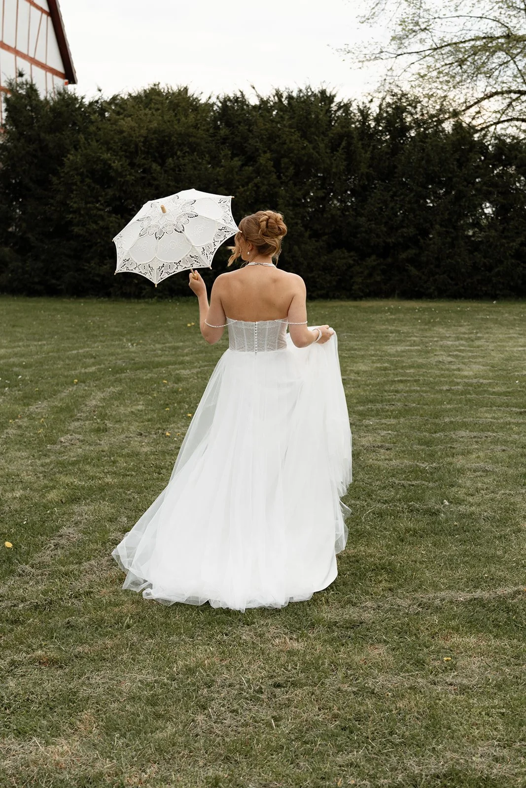 Eine Braut in weißem Hochzeitskleid mit einem weißen Parasol läuft über eine Wiese, im Hintergrund Bäume und ein Gebäude.