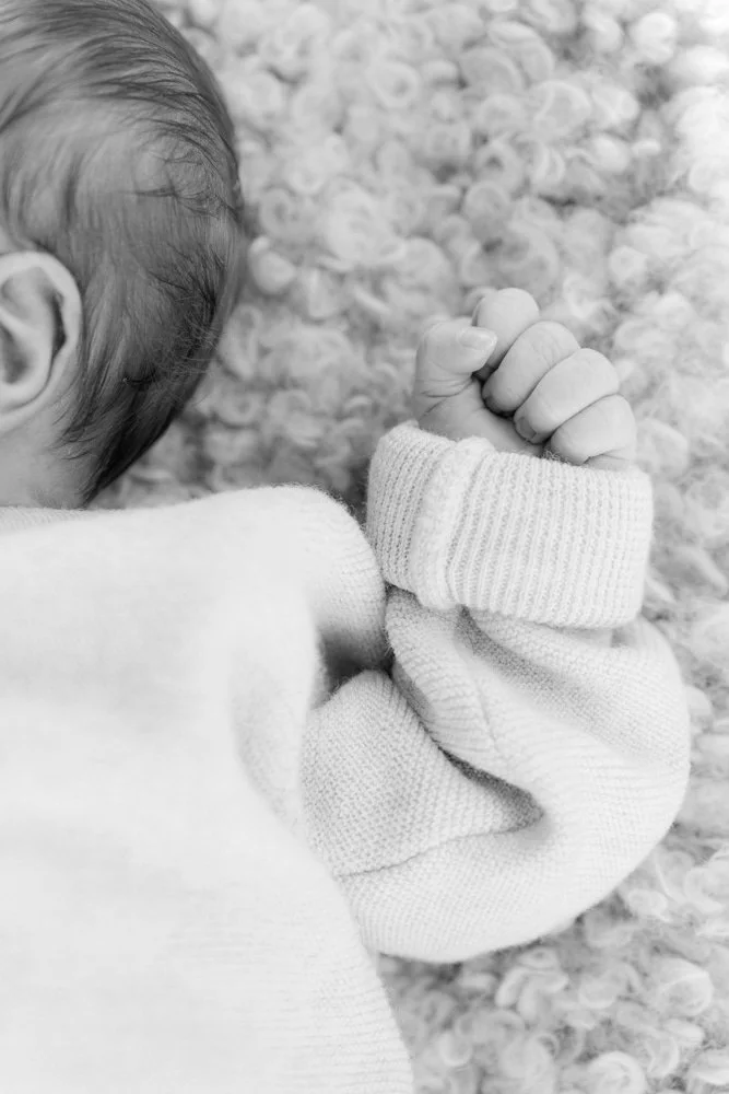 Nahaufnahme eines neugeborenen Babys, das auf einem weichen, gepunkteten Kissen liegt, mit den kleinen Hand umklammert. Das Bild ist schwarz-weiß.