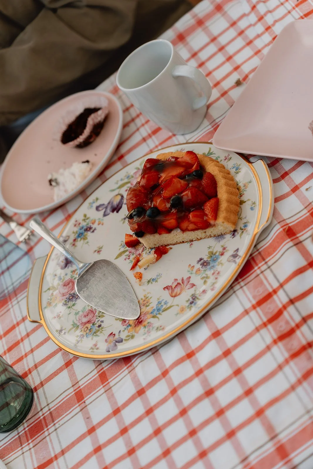 Blechkuchen mit Erdbeeren und schwarzen Beeren auf einem floralen Teller, ein halbleeres Geschirr mit Erdbeere und Schlagsahne, eine Tasse auf einem Tisch mit rot-weiß kariertem Tischtuch.