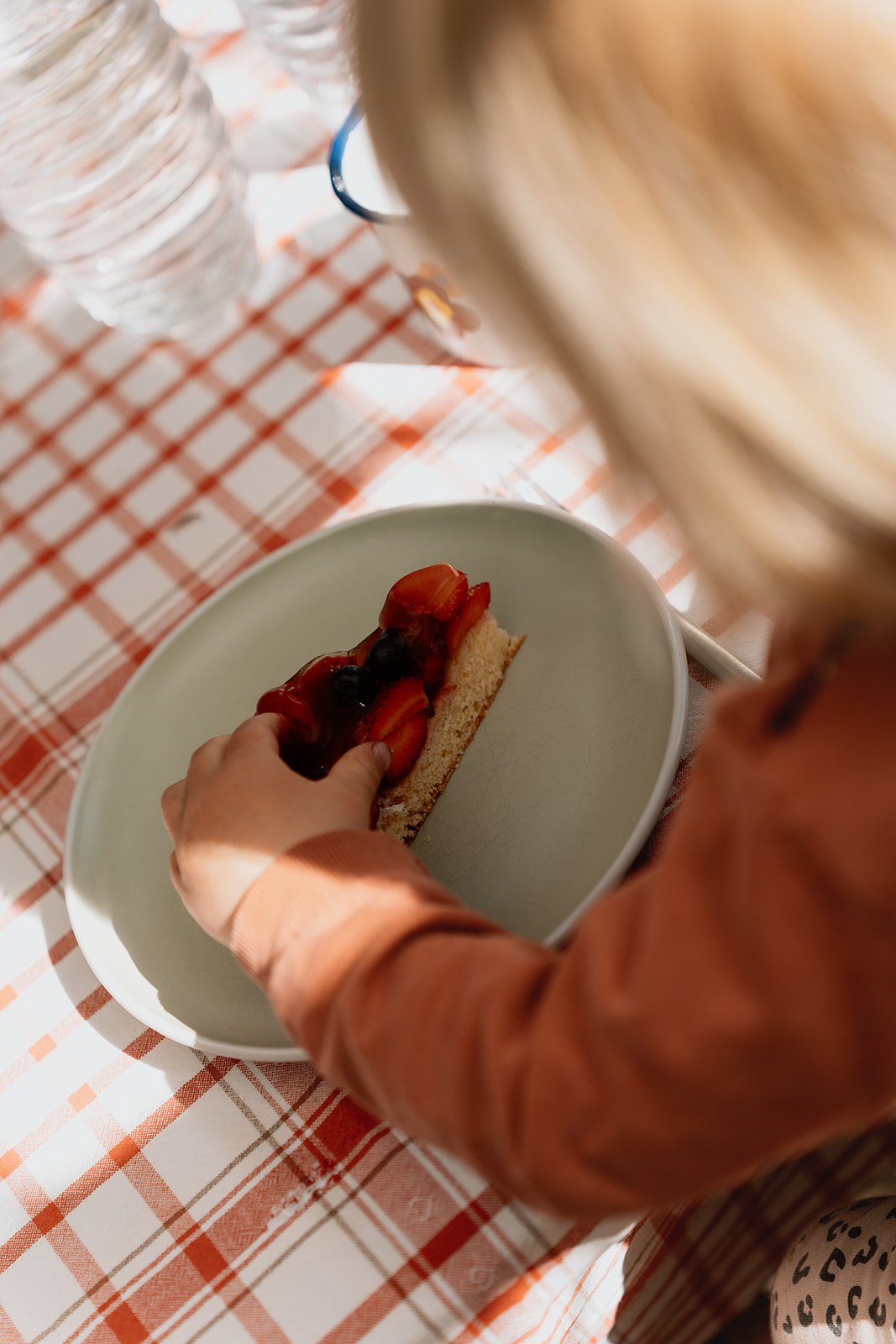 Ein Kind hält ein Stück Kuchen mit Erdbeeren und Heidelbeeren auf einem weißen Teller. Auf dem Tisch liegt eine rote kariert Tischdecke, und im Hintergrund sind Wasserflaschen sichtbar.