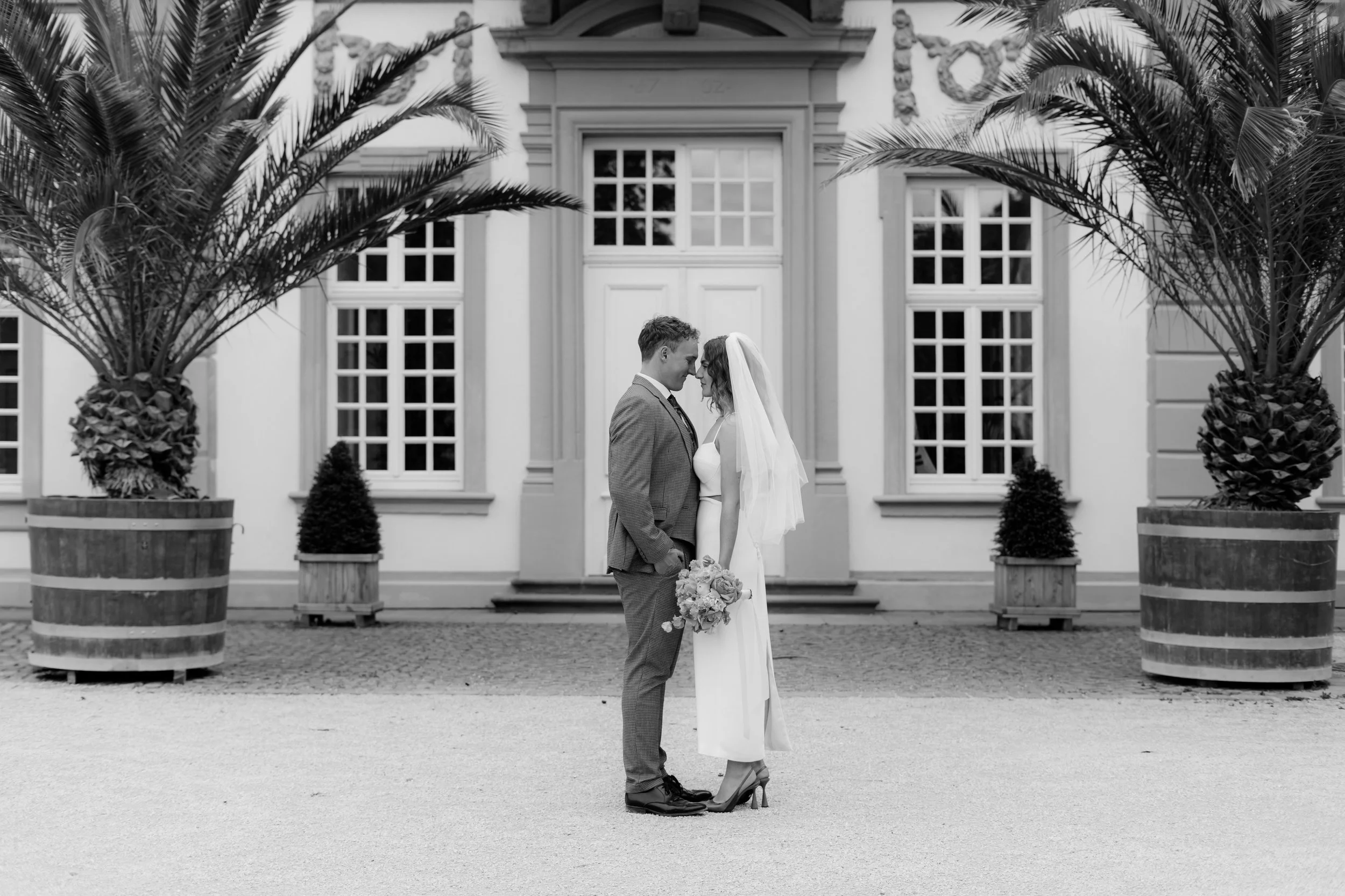 Ein Brautpaar steht sich vor einem weißen Gebäude im schwarzen und weißen Foto, umarmt sich und blickt sich in die Augen. Die Braut hält einen Blumenstrauß, trägt ein langes Kleid mit Schleier, der Bräutigam trägt einen Anzug.