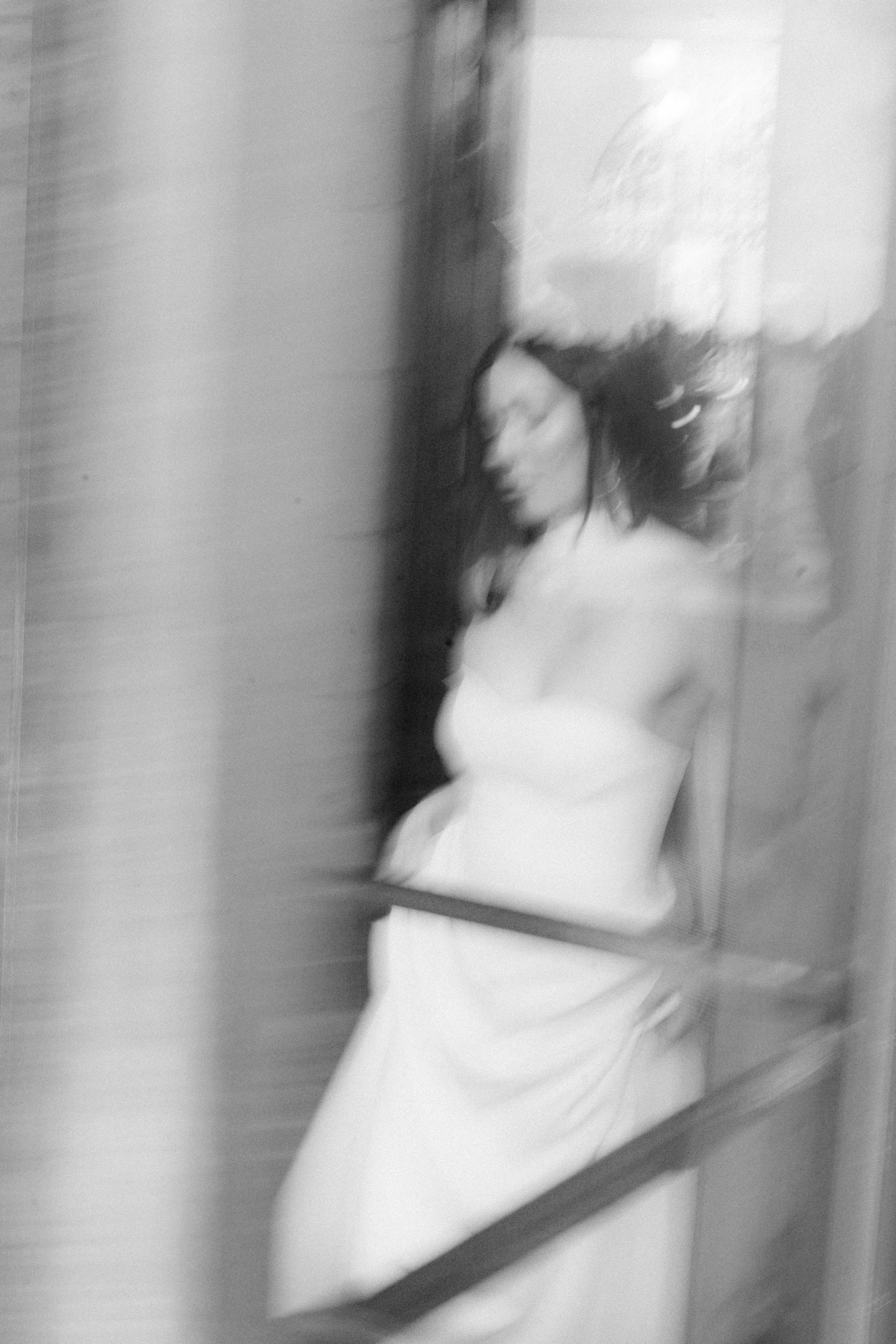 Schwarz-Weiß-Foto einer schlafenden Frau in einem weißen Kleid, aufgenommen durch eine reflektierende Glasscheibe, leicht verwackelt.