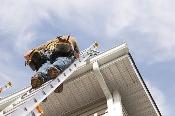 Technician cleaning residential gutters from ladder near downspout