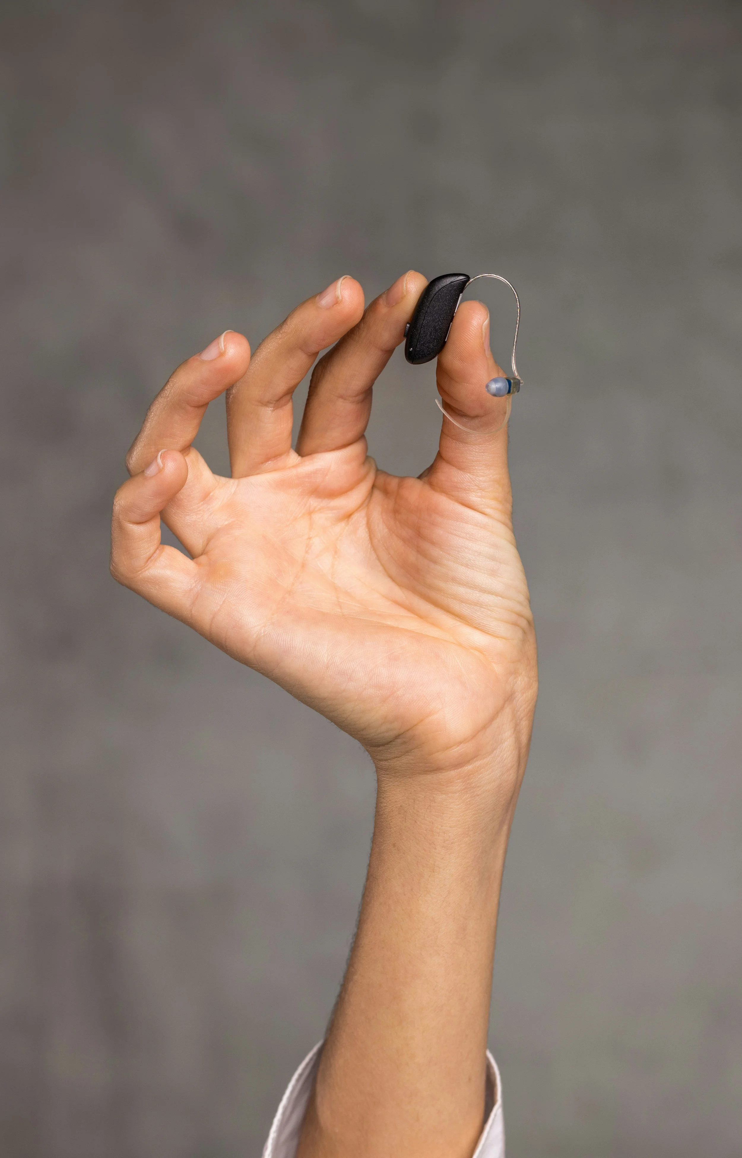 Close-up of a hand holding a small, bluetooth, behind-the-ear (receiver-in-the-ear) hearing aid against a gray background.