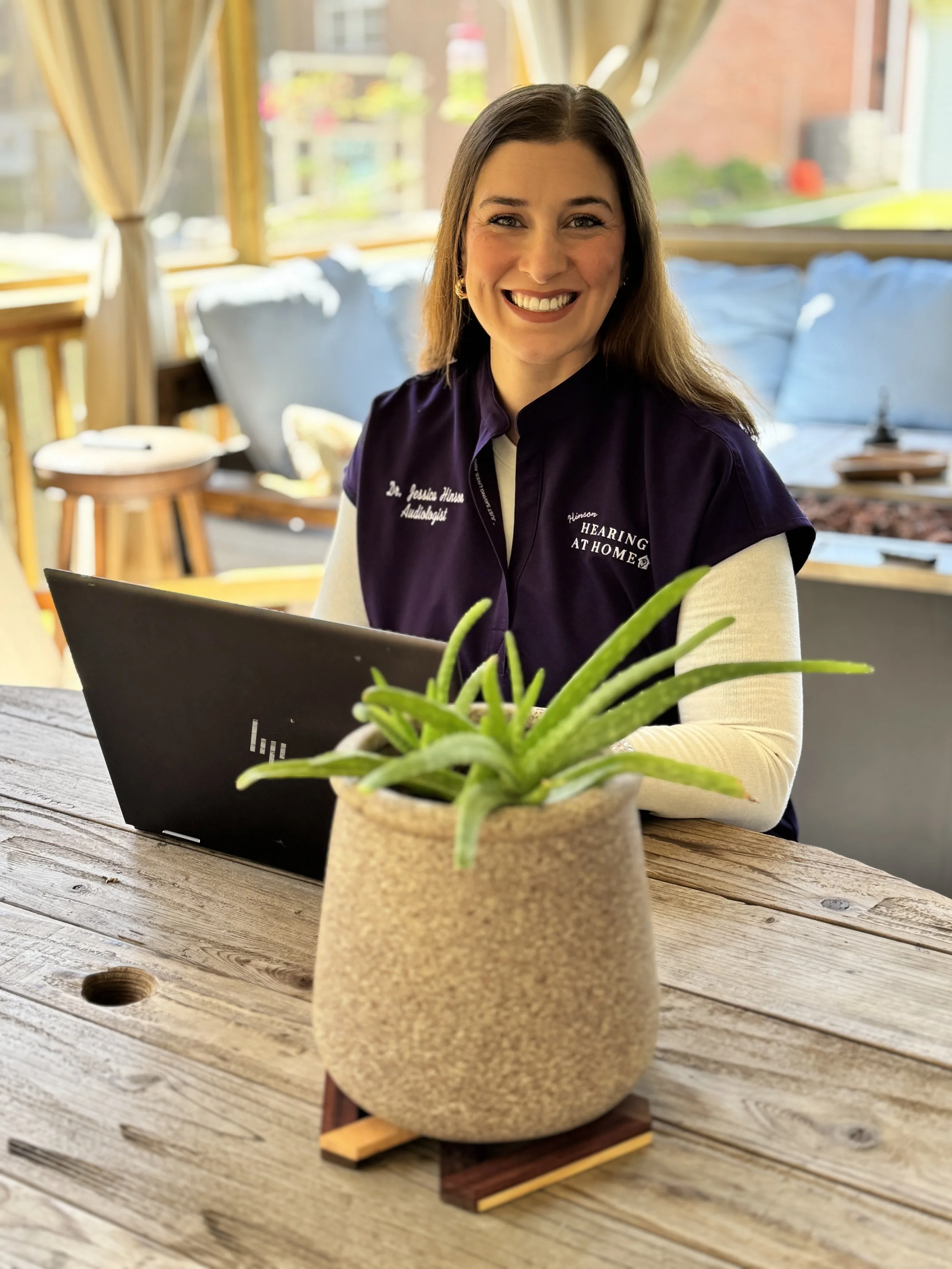 An audiologist working on a computer on the back porch of a patient's home with sun shining and a plant in the foreground.