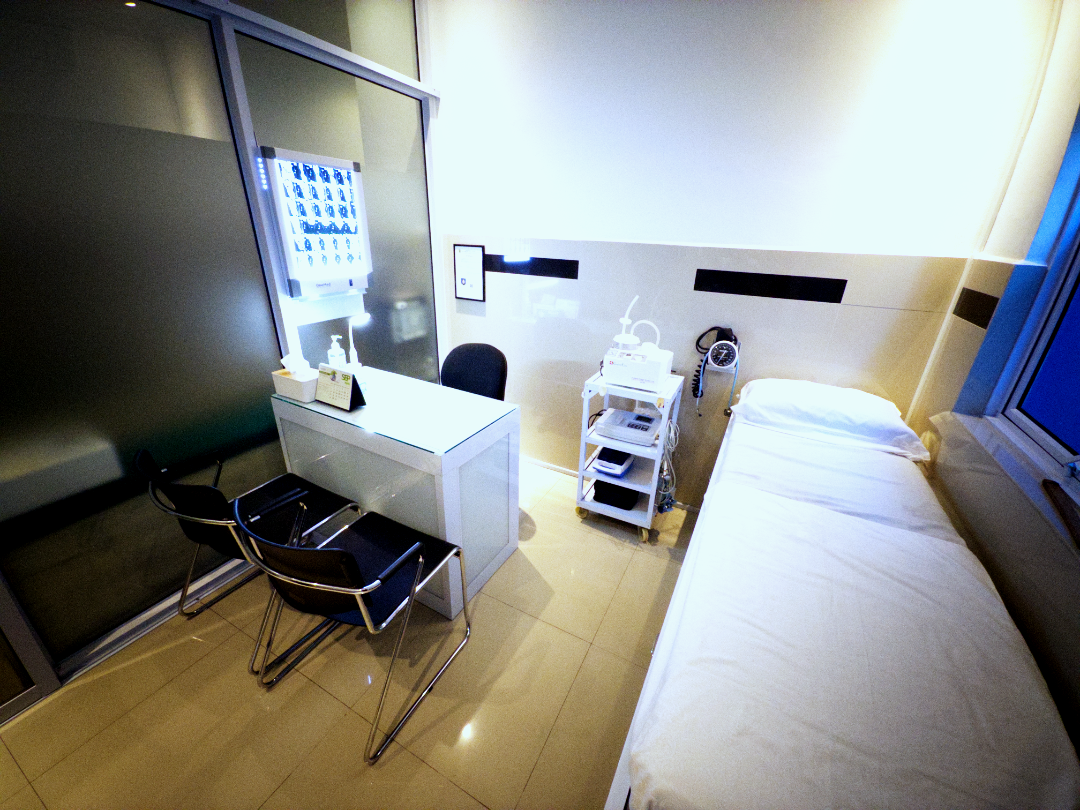 A hospital room with a bed, medical equipment, a white desk with two chairs, and a wall-mounted monitor.