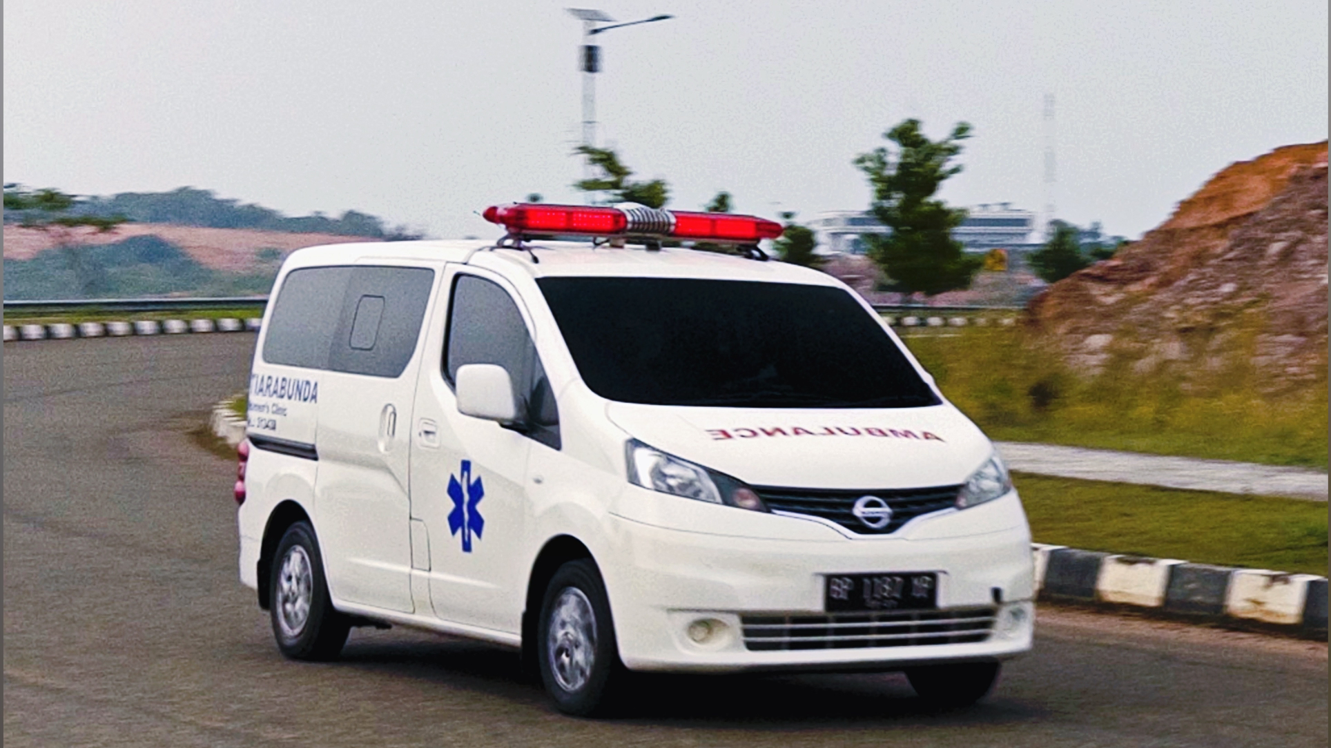 A white ambulance van with blue medical emblem and red emergency lights on top, driving on a curved road with a rocky landscape and trees in the background.