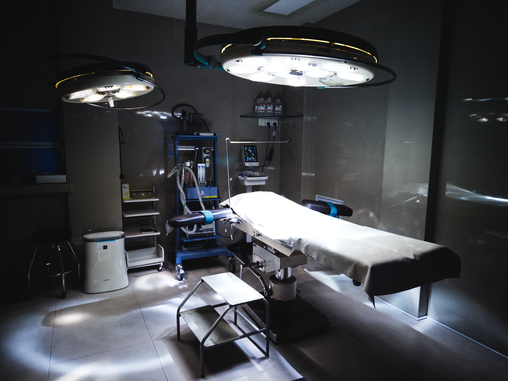 Empty operating room with surgical lights, patient bed with white sheets, medical equipment, and dark surroundings.