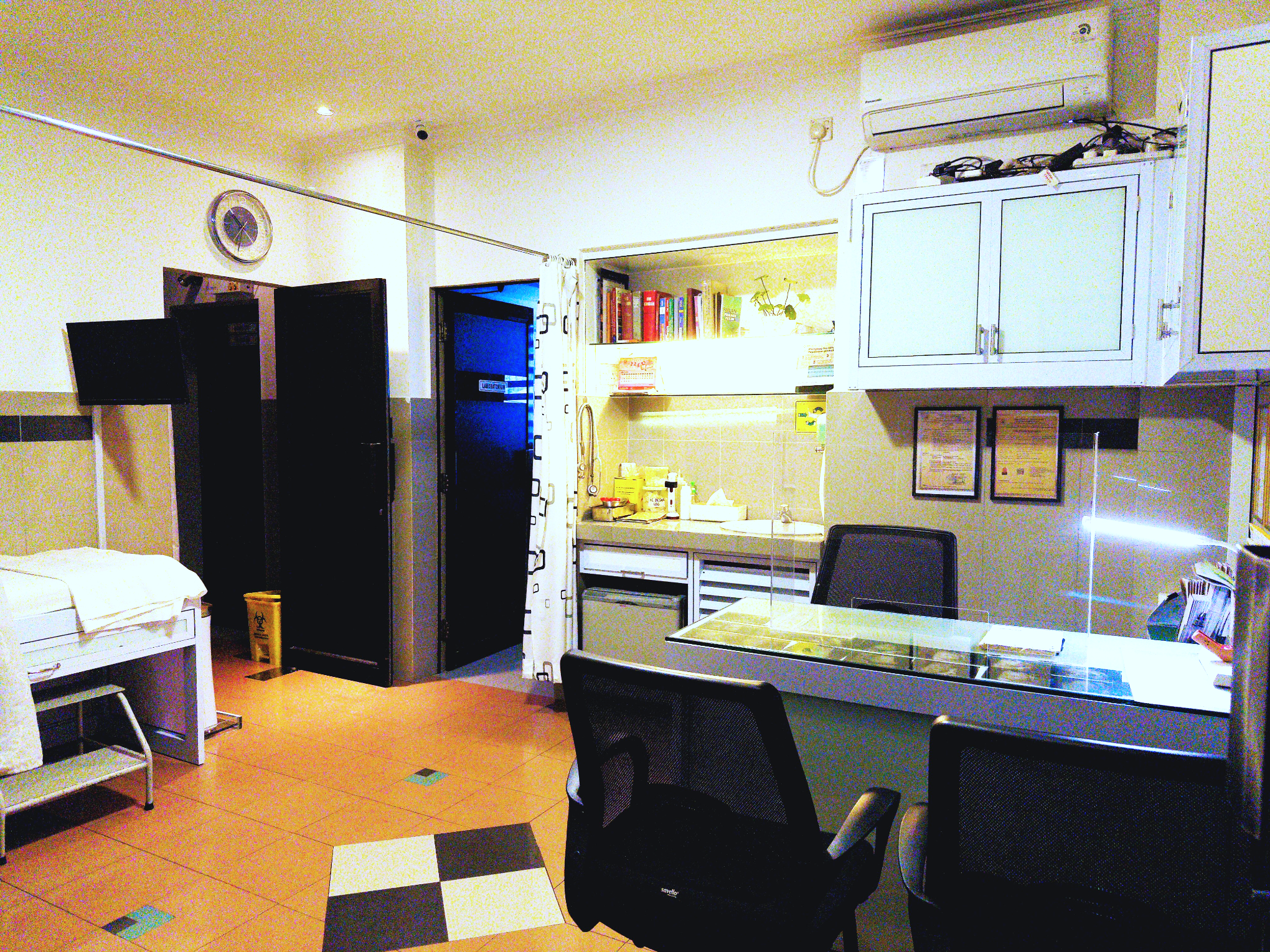 Small room with a hospital bed, a black room divider, a small kitchenette with yellow items, a white desk with chairs, and an air conditioning unit on the wall.
