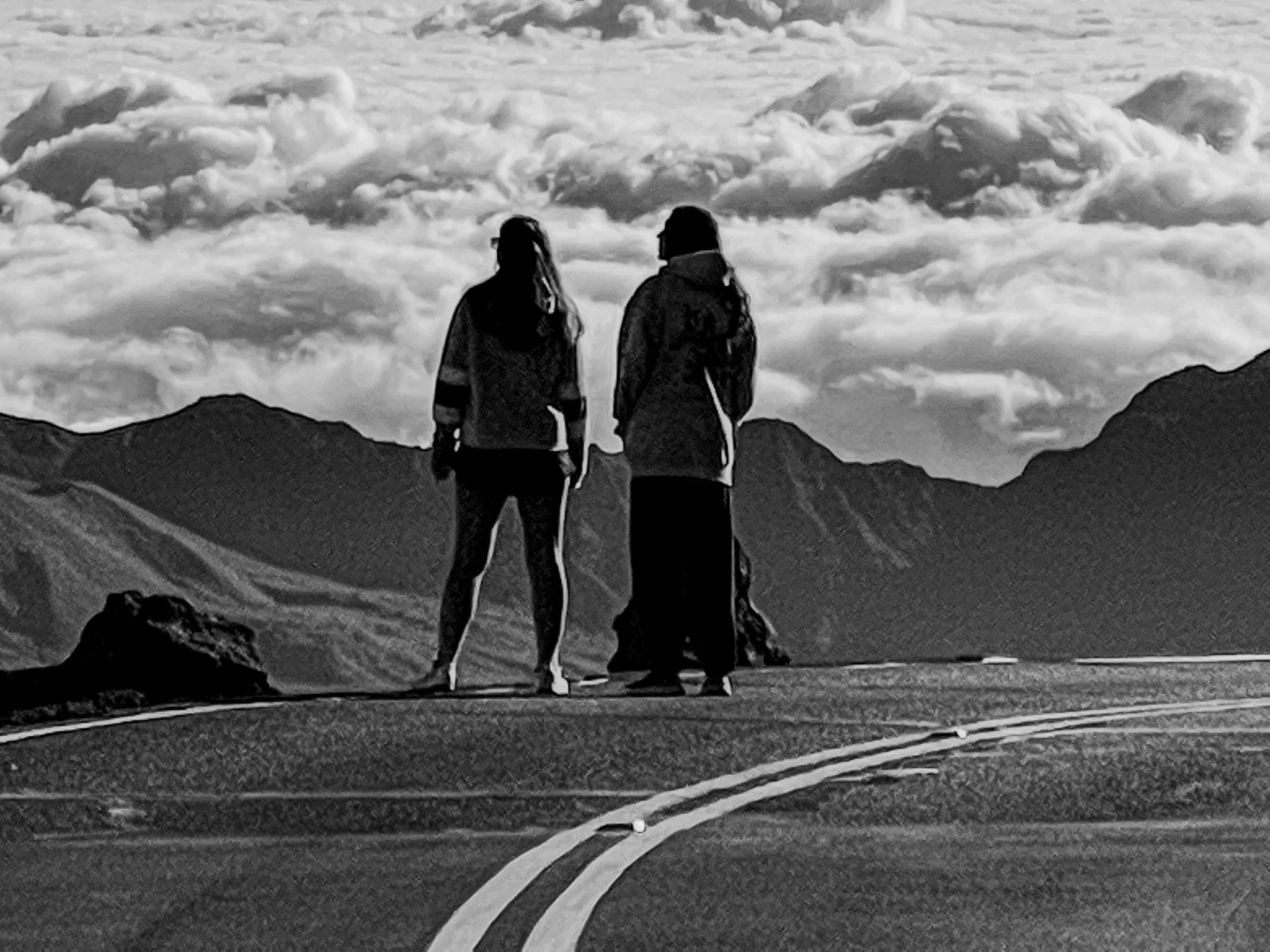Two women stand on a winding mountain road, looking at the distant mountains and clouds in the sky, in black and white.