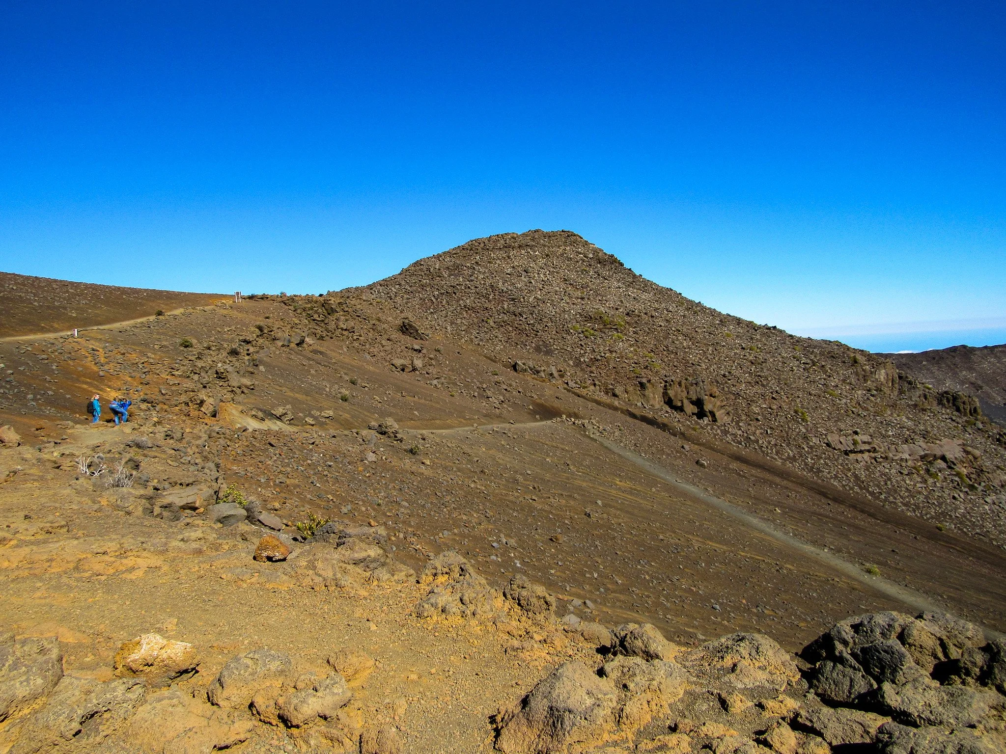 Two hikers in blue jackets walking along a rocky mountain trail with a large hill in the background under a clear blue sky.
