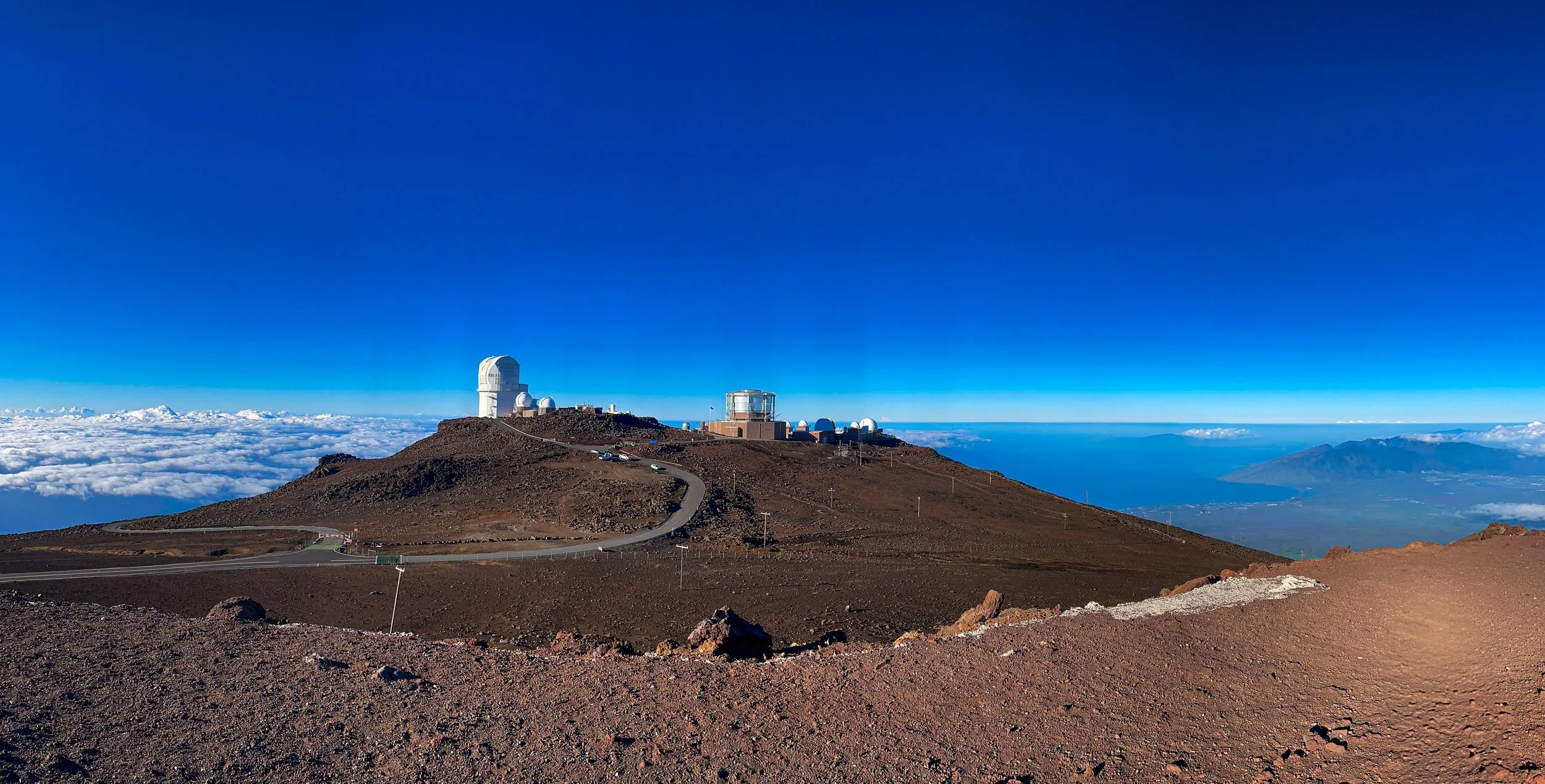 View of a mountain summit with a clear blue sky, featuring several observatory buildings, including a large white dome, atop the rugged, barren terrain.