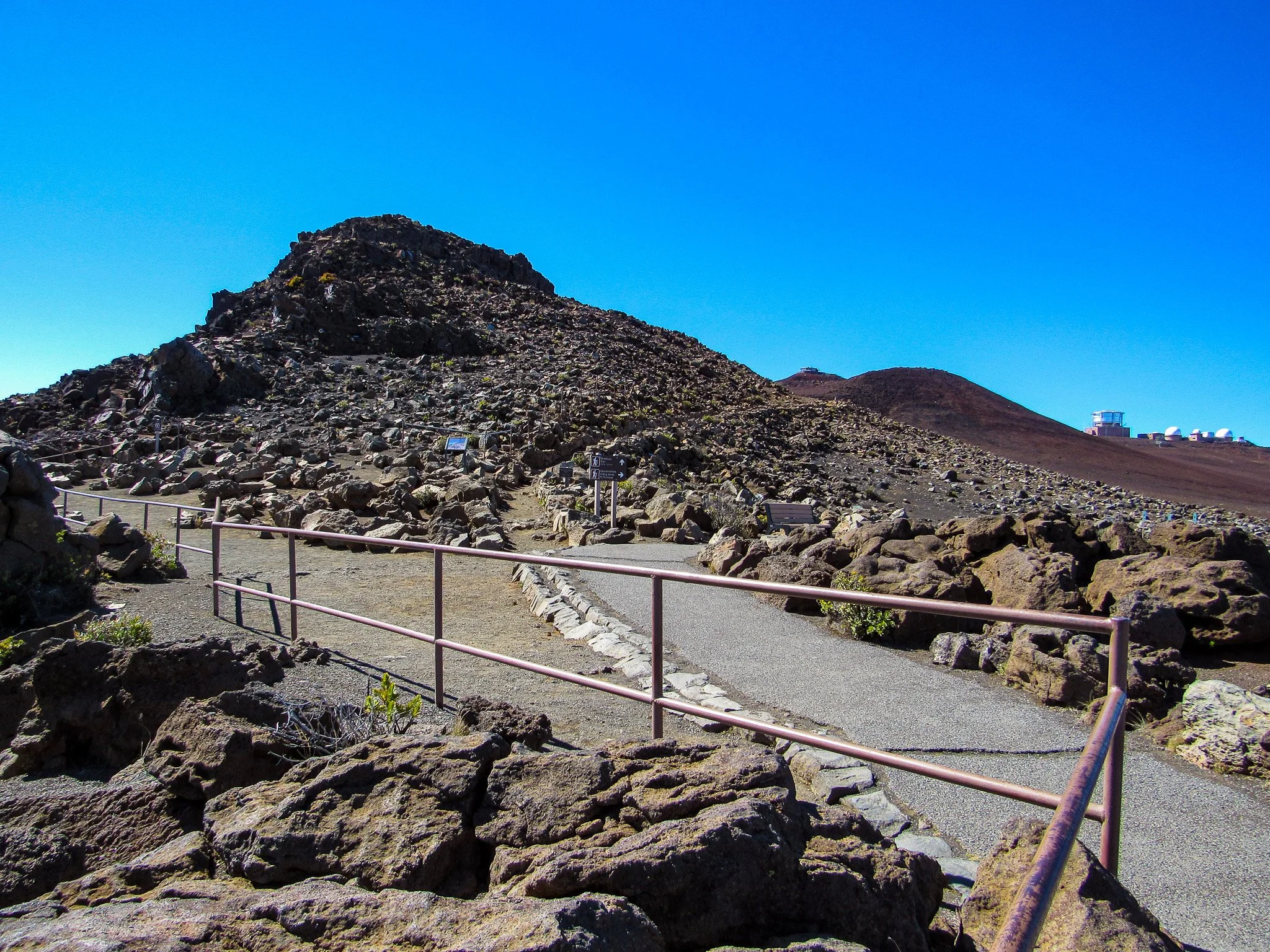 A rocky volcanic landscape with a dirt pathway and metal railing leading up a volcano under a clear blue sky. Observatory buildings are visible on the horizon.