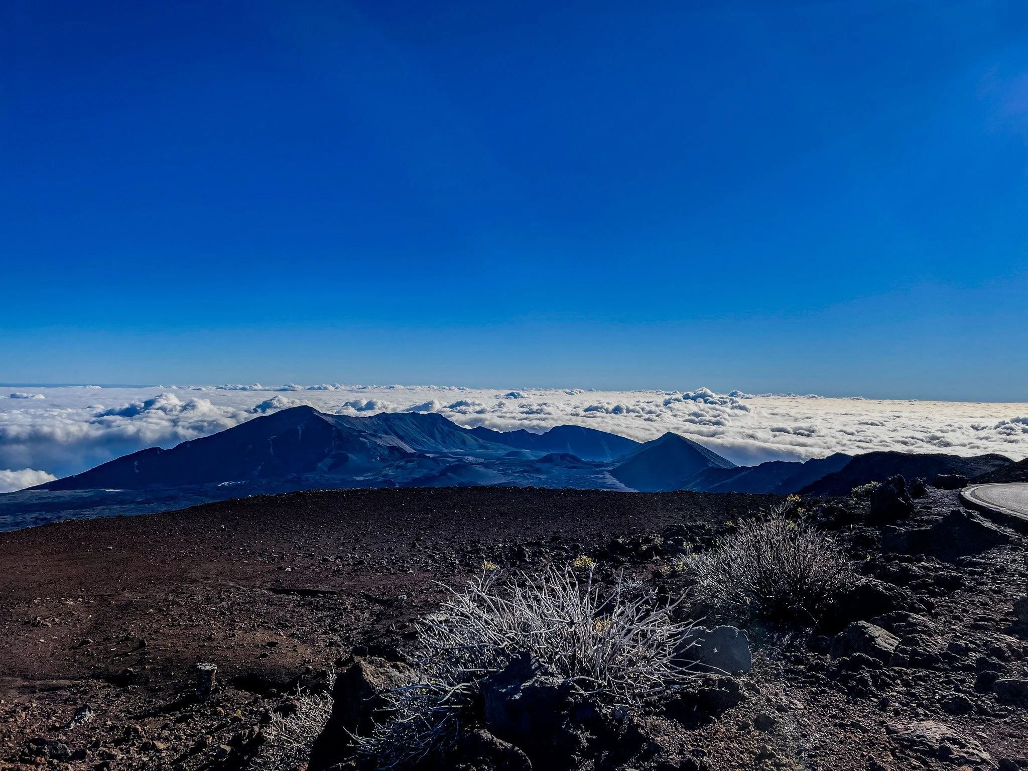 A mountainous landscape beneath a clear blue sky with clouds below the mountain peaks and dry, rocky terrain in the foreground.