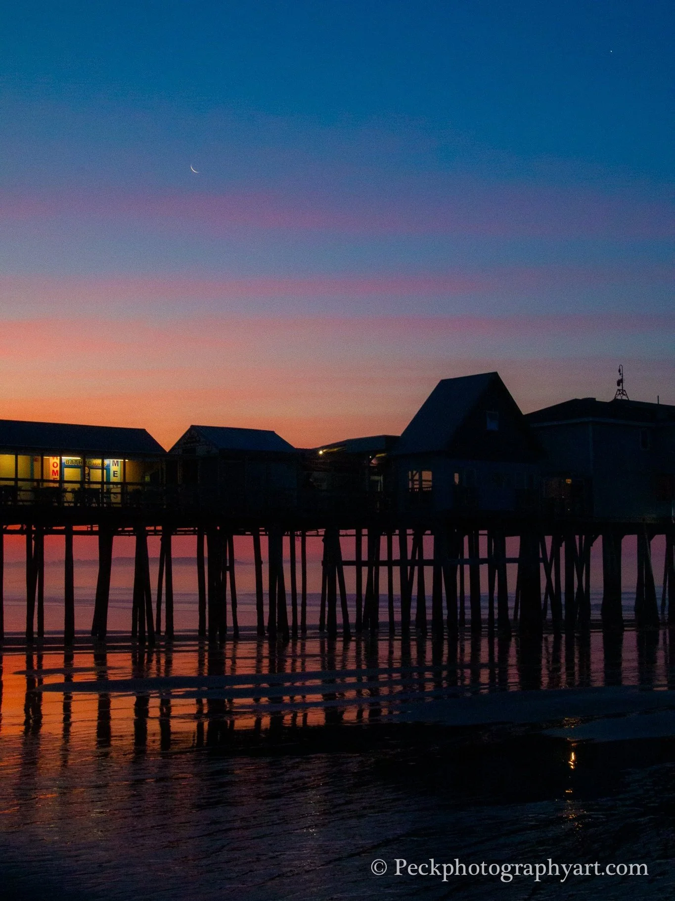 Sunset over a pier with silhouetted buildings and a crescent moon in the sky.