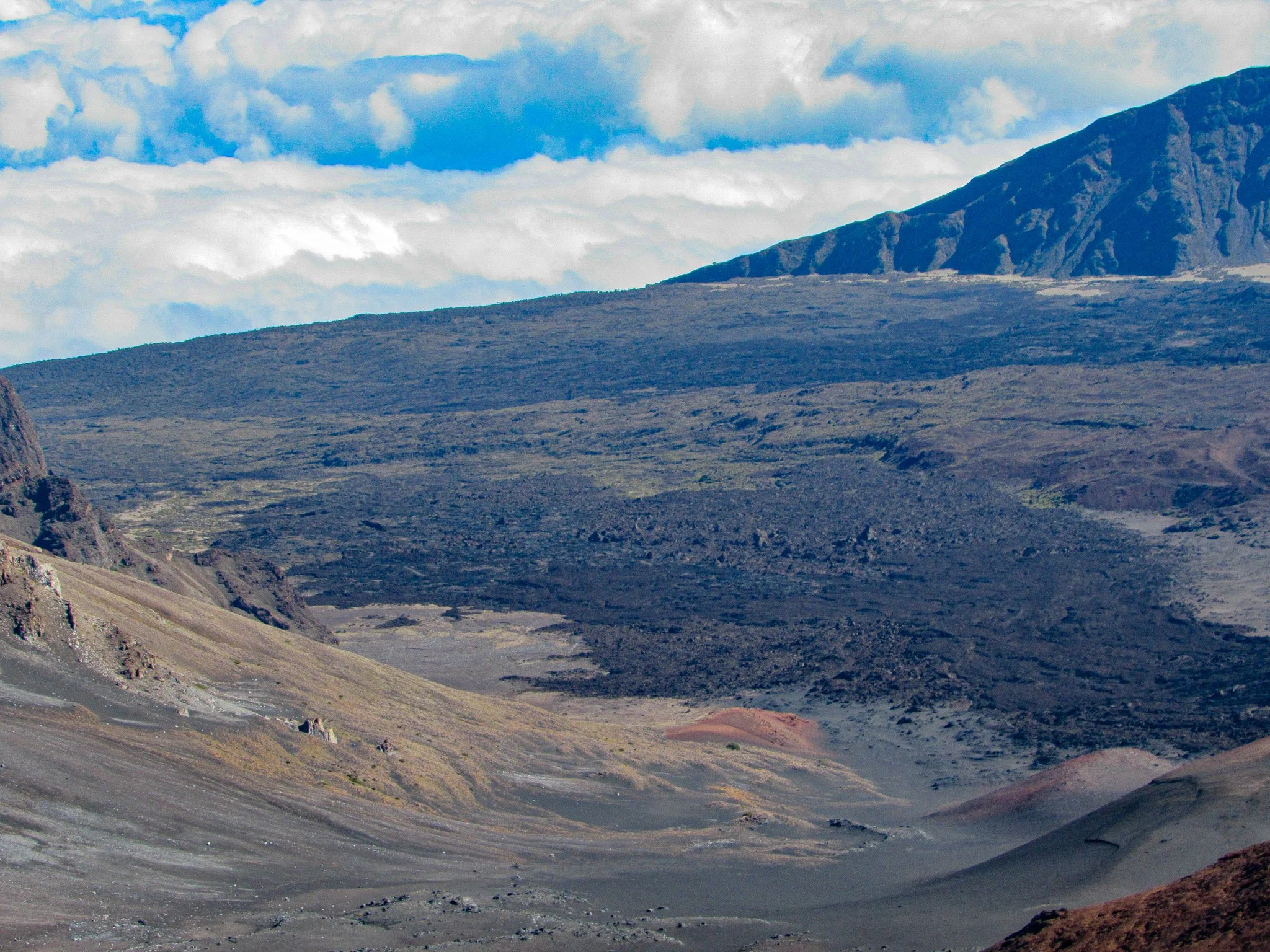 A volcanic landscape with dark, rugged terrain and a mountain in the background under a partly cloudy sky.