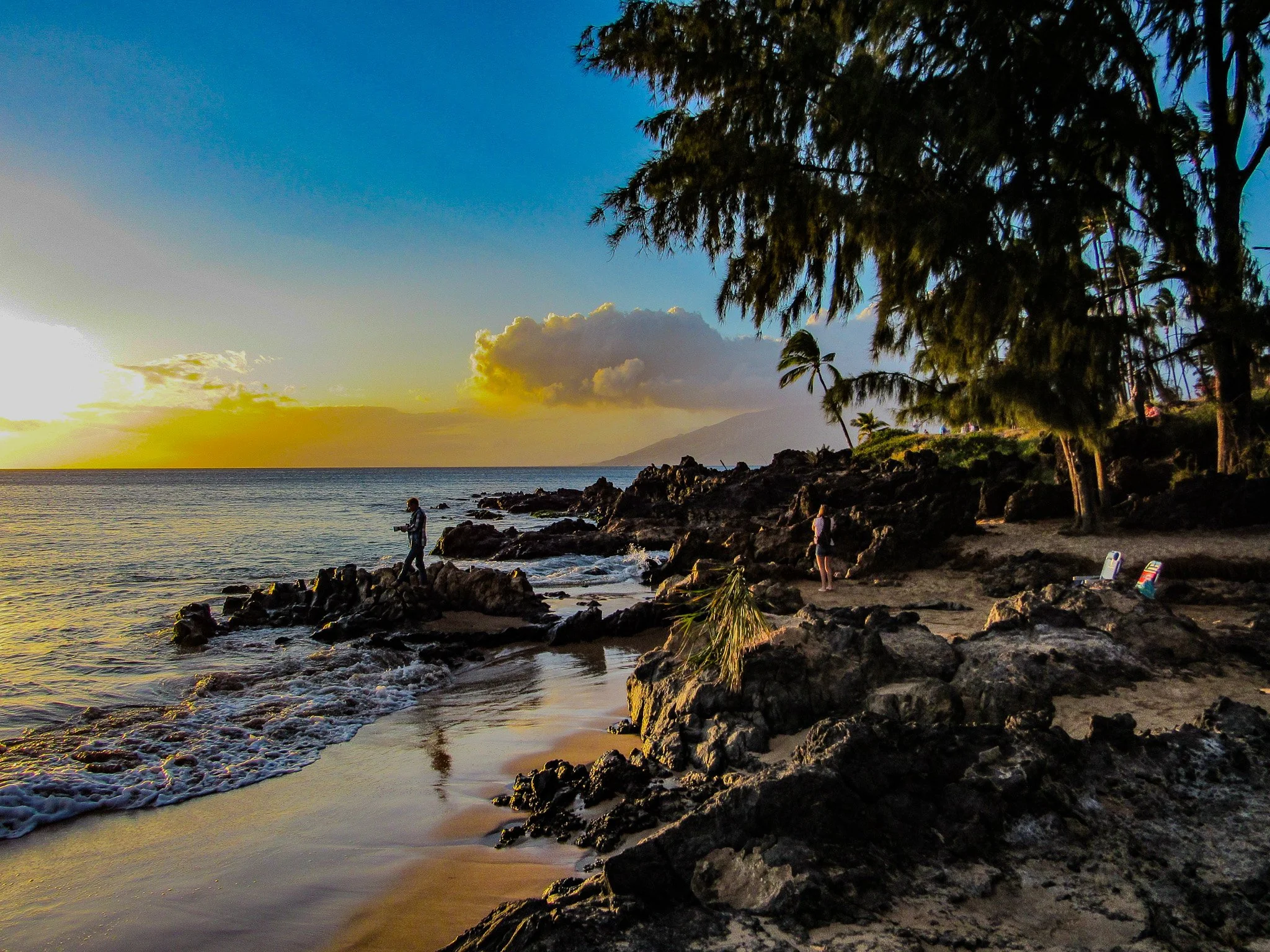 Sunset along the Kihei coastline on Maui with fisherman standing on volcanic rocks and palm trees overlooking the Pacific.