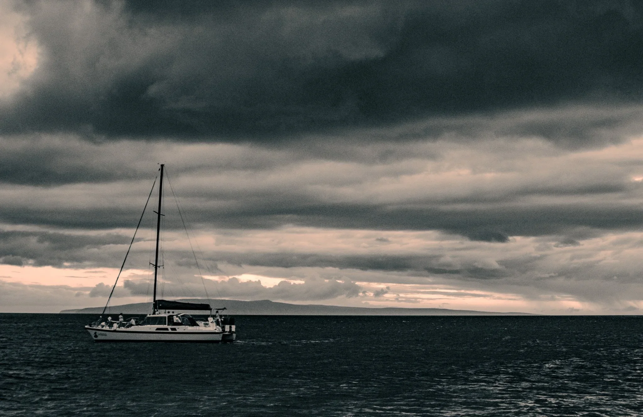 Black and white sailboat on the Maui ocean beneath layered storm clouds with dark water and distant island horizon