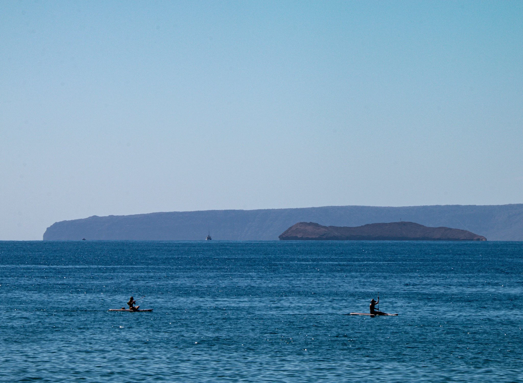 Distant view of Molokini Crater rising from the Pacific Ocean between Maui and the island of Kahoʻolawe with paddle boarders off the coast of Kihei.
