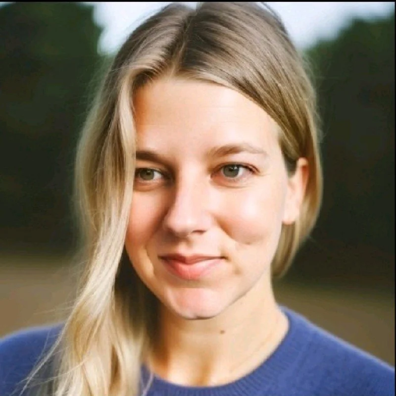 A young woman with blonde hair and fair skin smiling outdoors, wearing a blue sweater, with blurred trees in the background.