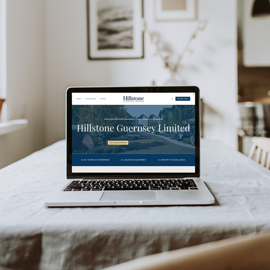 Laptop on a dining table displaying the Hillstone Guernsey Limited website, with framed artwork and a bouquet of flowers in the background in a cozy, well-lit room.