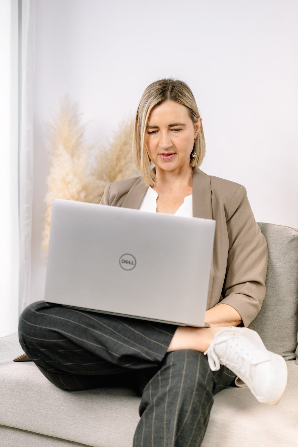 Woman sitting on grey couch with legs crossed, using a silver Dell laptop, with a beige blazer, white top, pinstriped pants, and white sneakers, in a room with a white wall and decorative plant in the background.