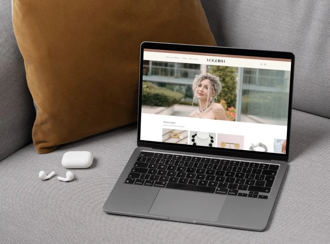 A laptop on a grey sofa next to a pair of wireless earbuds and their charging case, with a brown pillow in the background.