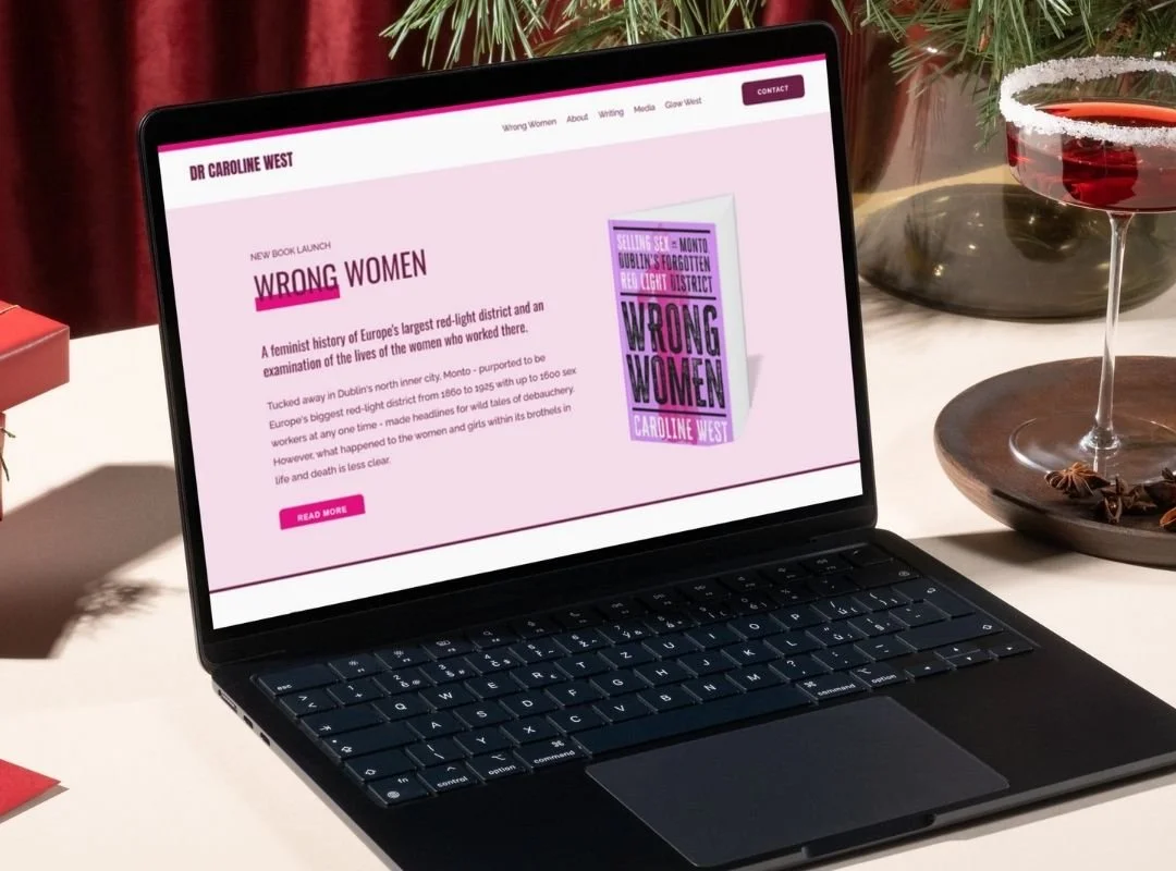 A black laptop sitting on a white table with red curtains in the background and a martii glass filled with a red drink bedside it. The laptop screen has Dr Caroline West's website