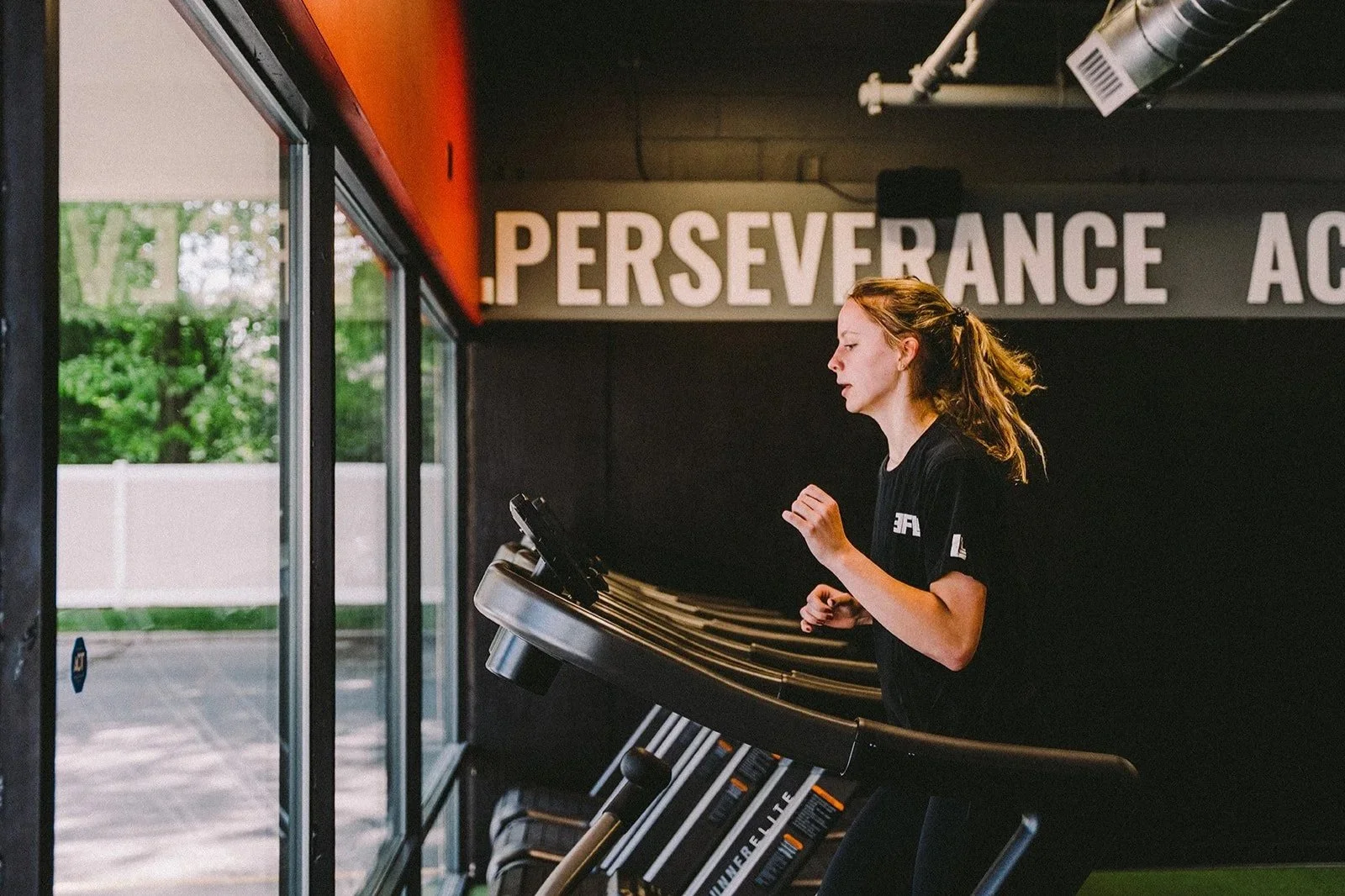 A woman jogging on a treadmill in a gym with a large window showing outdoor greenery. The gym wall behind her has the words 'PERSEVERANCE' and 'AC' visible.