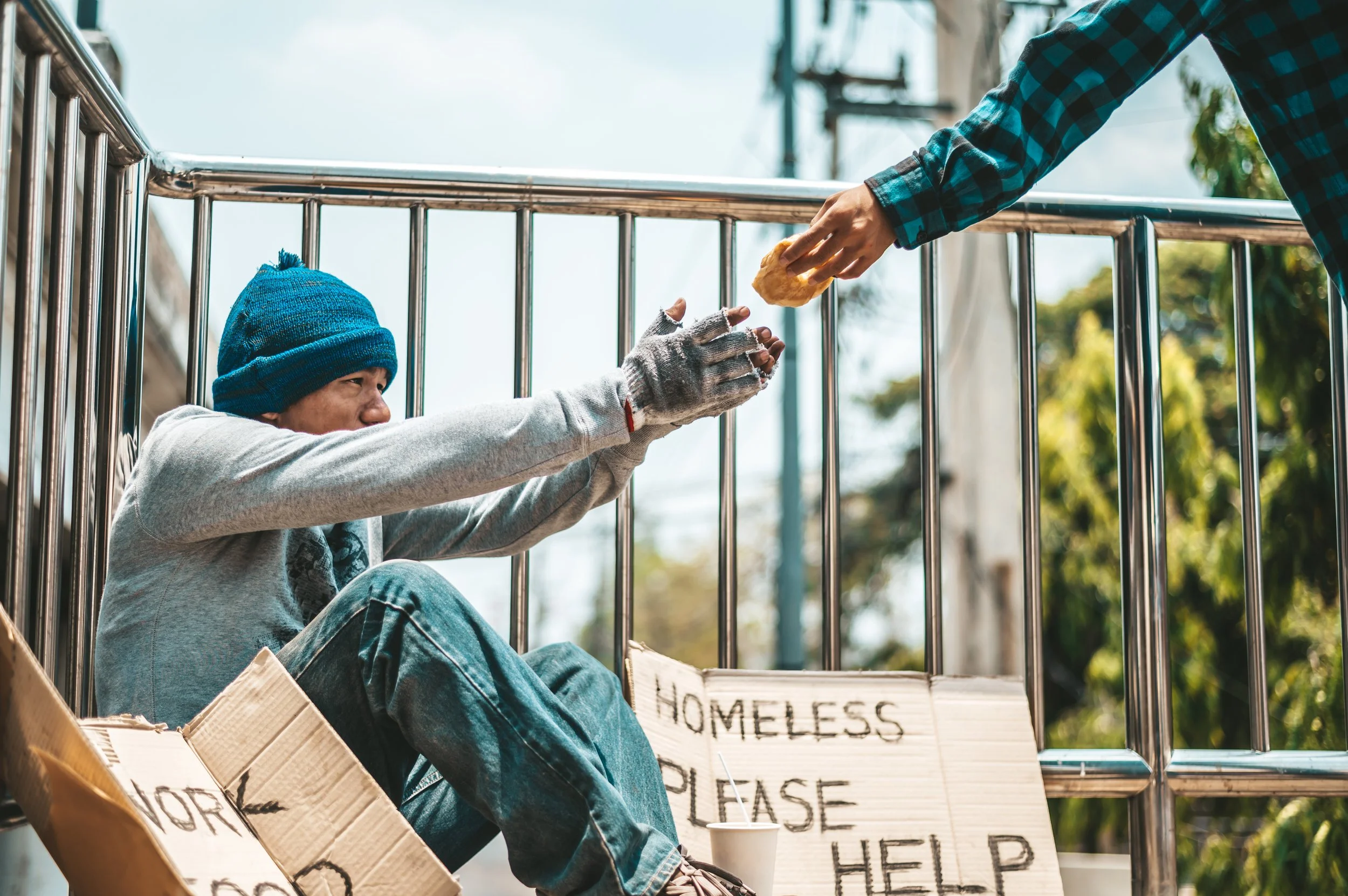 A person experiencing homelessness sitting on the ground in a cardboard shelter, wearing a blue beanie and gray hoodie, reaching out to a passerby for help by accepting food. A sign reading 'HOMELESS PLEASE HELP' is propped against the shelter.