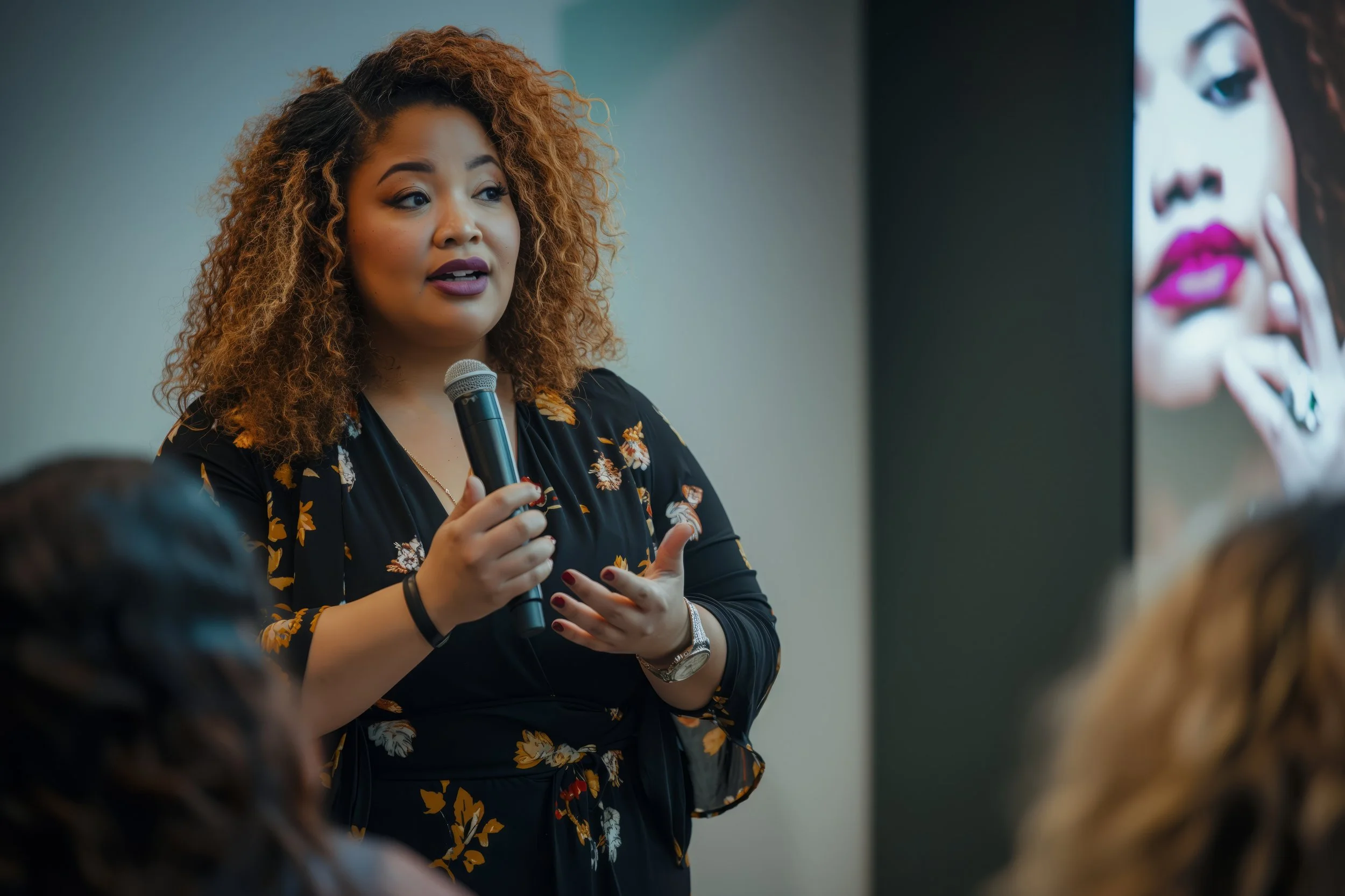 A woman with curly hair is speaking into a microphone during a presentation, with a large image of a woman with makeup displayed on a screen next to her.