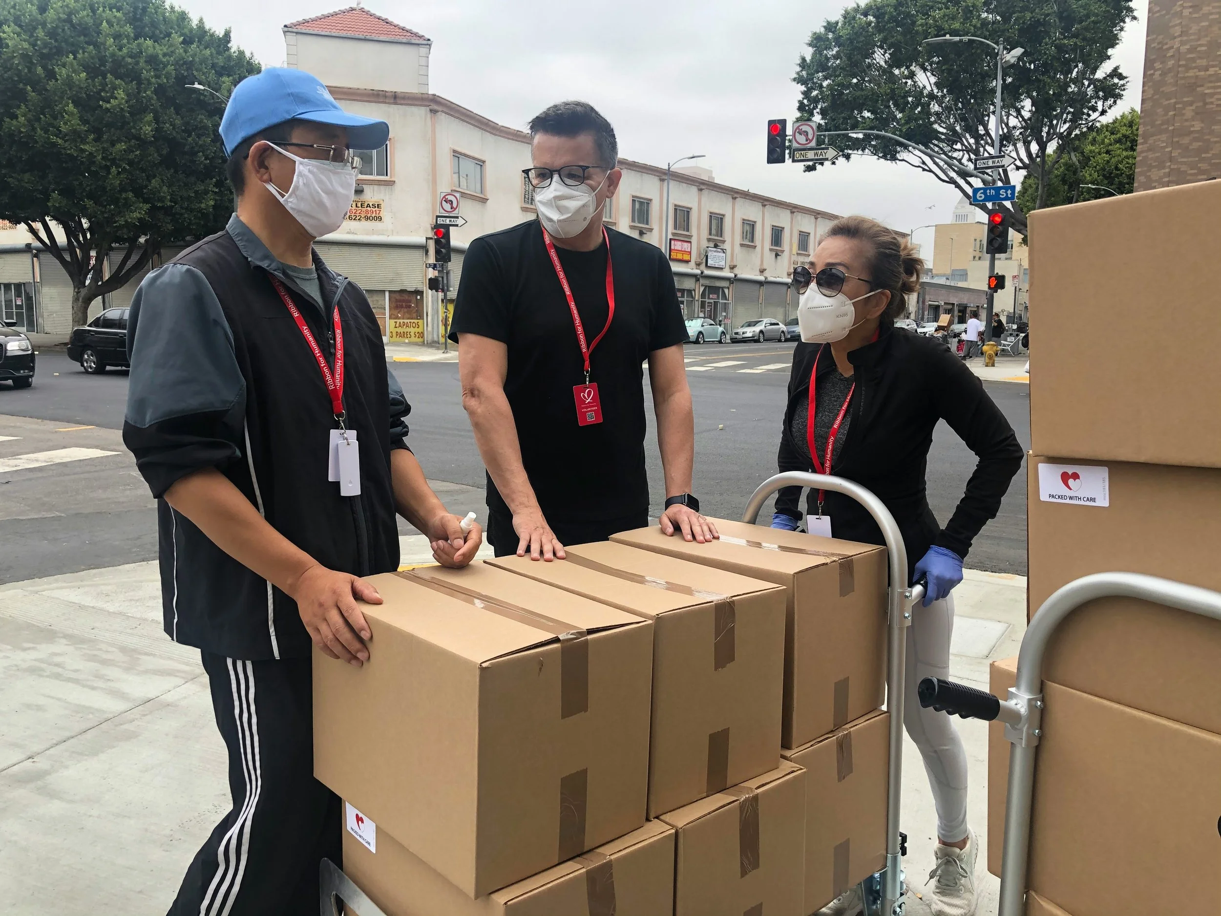 Three people wearing masks and gloves with boxes on a cart on a city sidewalk.