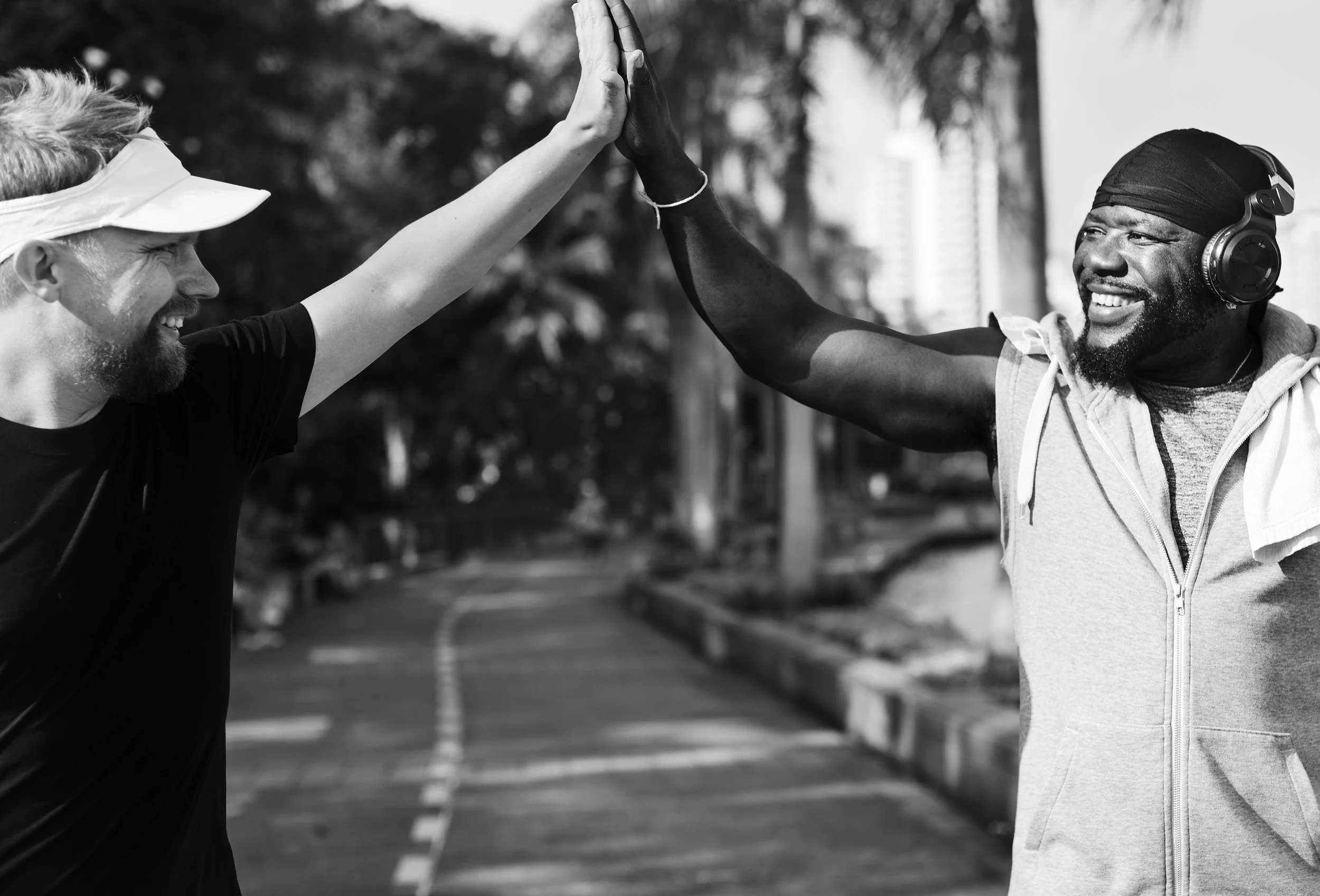 Two men high-fiving during a workout on a city street, one in a black t-shirt and a visor, the other in a sleeveless hoodie and headphones, both smiling.