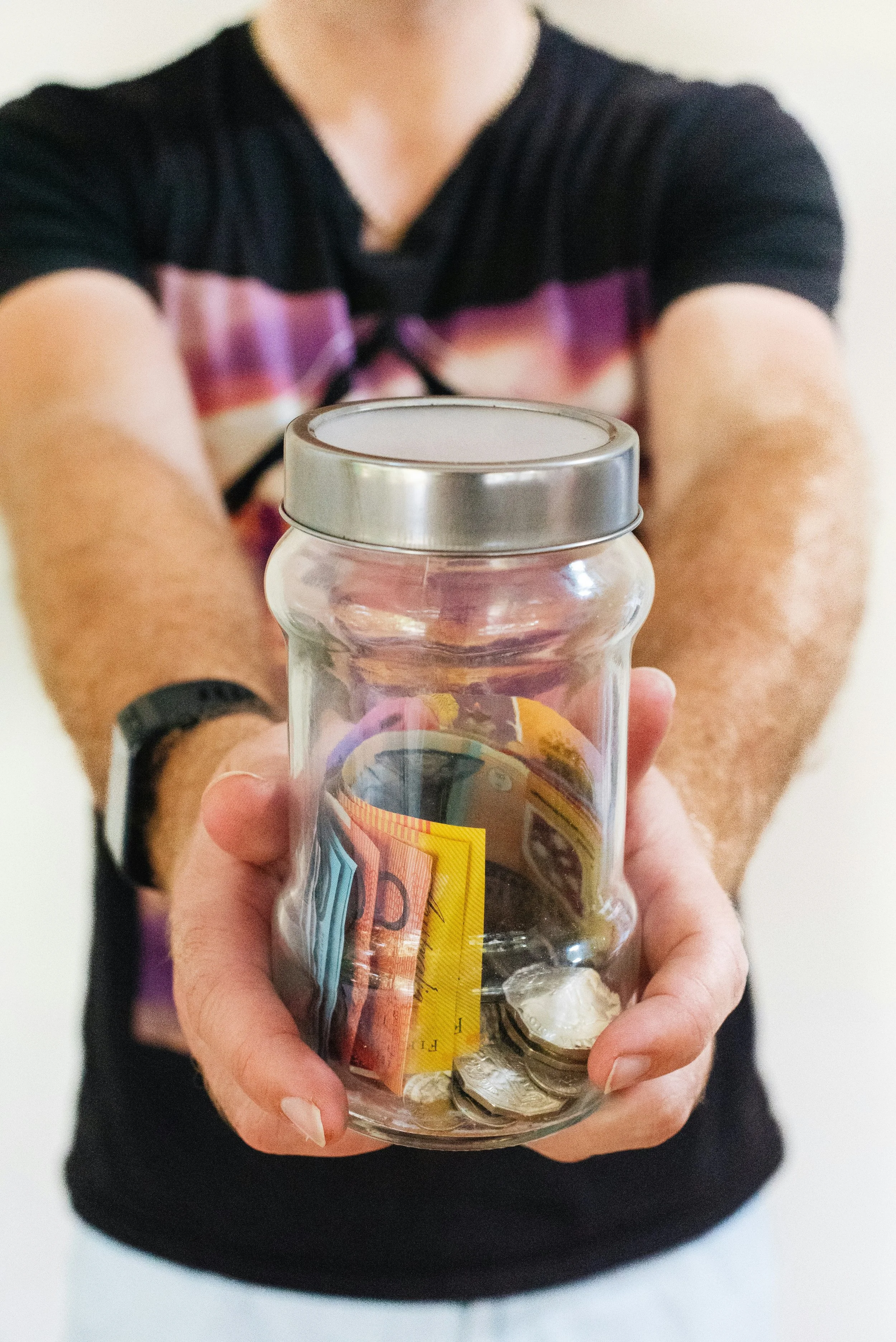 Person holding a glass jar with cash and coins inside, extending it towards the camera.