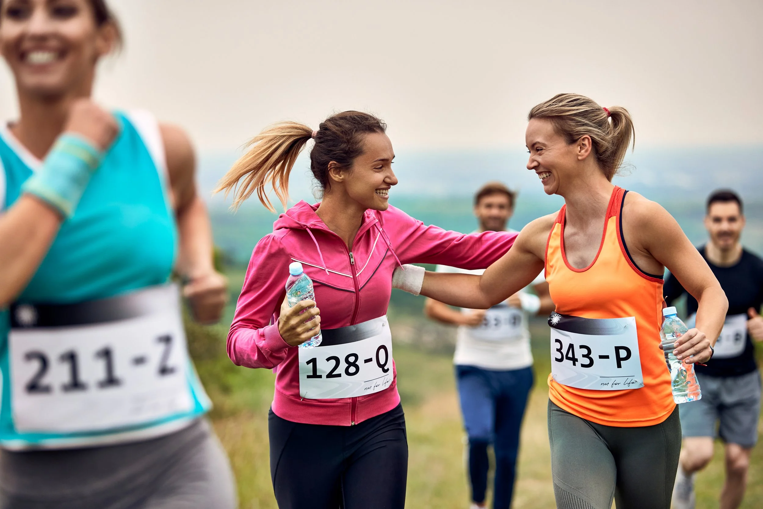 Two women smiling and celebrating during a marathon race, wearing race bibs and holding water bottles, with other runners in the background on an outdoor trail.