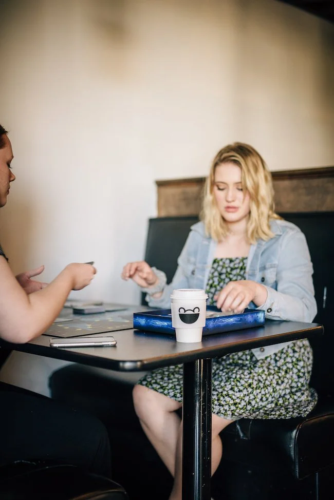 A woman with blonde hair sitting at a table with a coffee cup that the Ready Teddy's logo, while another person, partially visible, is interacting with her.