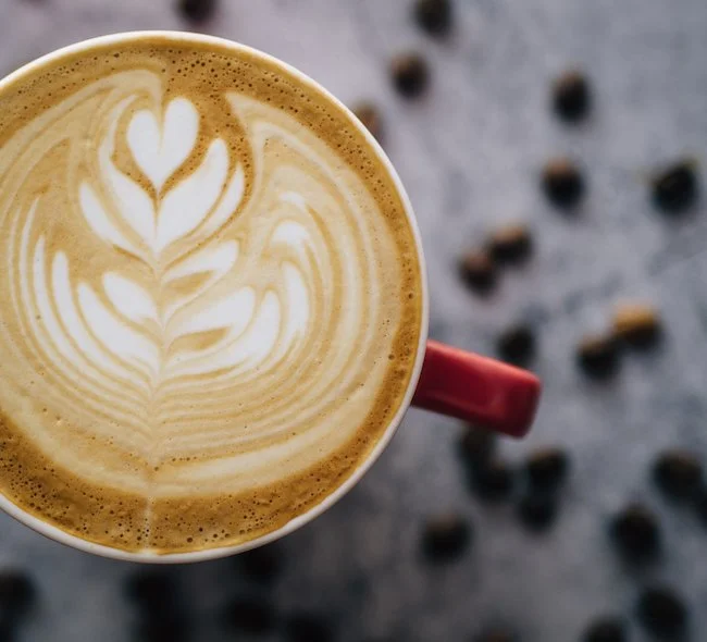 A top view of a latte with latte art in the shape of a heart and leaf, served in a red mug with coffee beans scattered around.