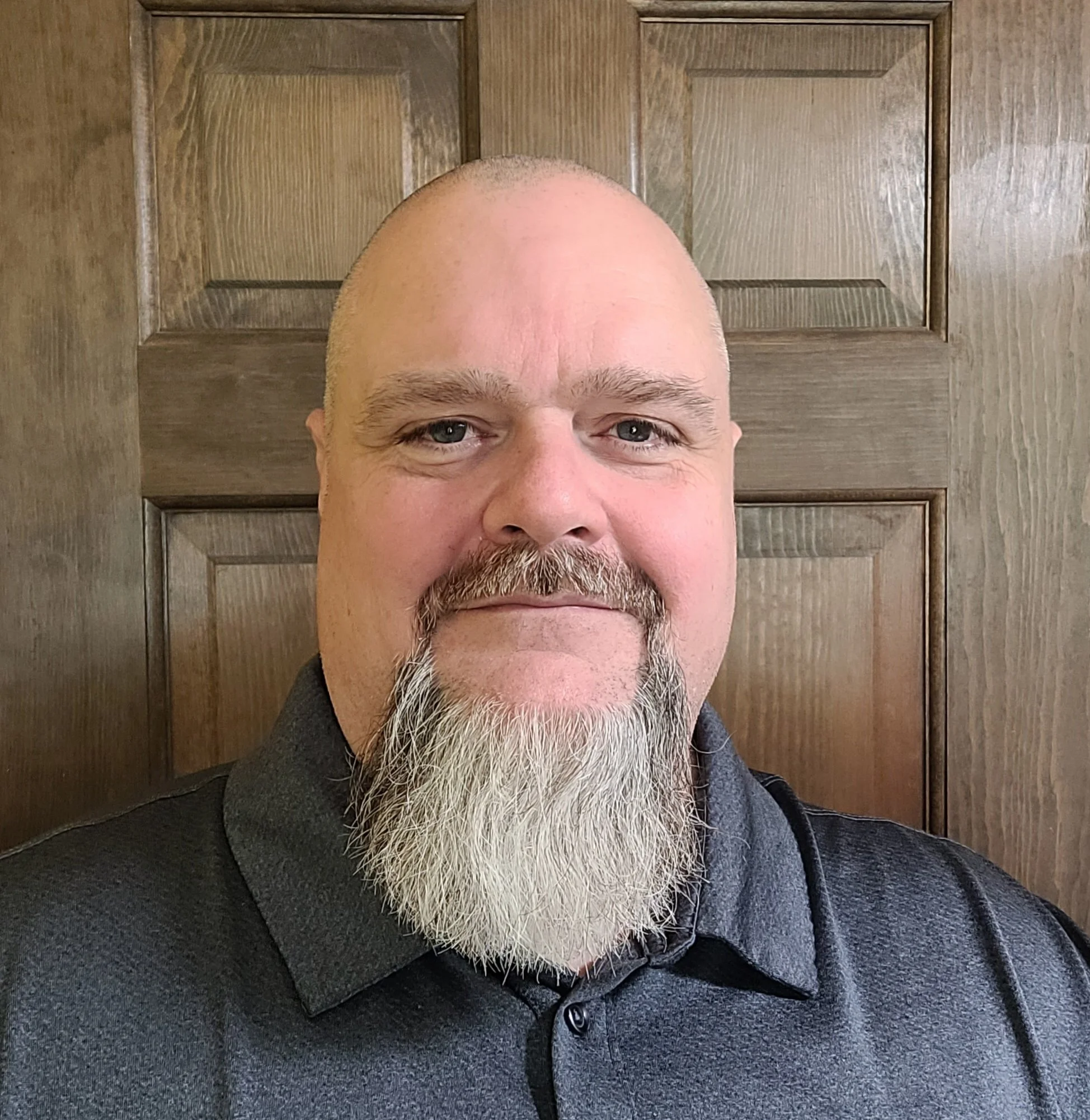 A man with a bald head and gray beard taking a selfie in front of a wooden door with panels.