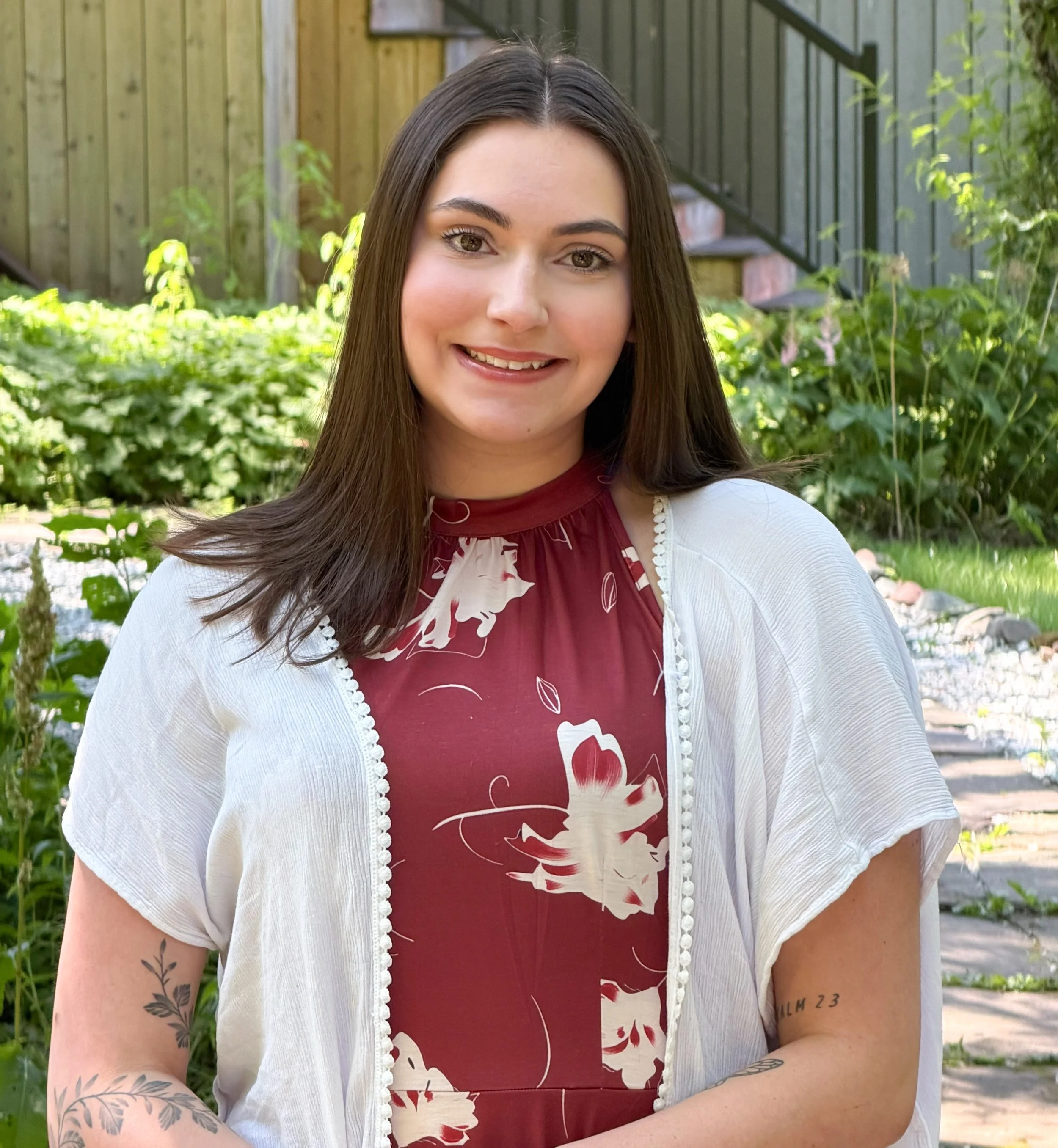 A young woman with long dark hair and tattoos on her arm smiling outdoors in front of a garden with green foliage and a wooden fence.
