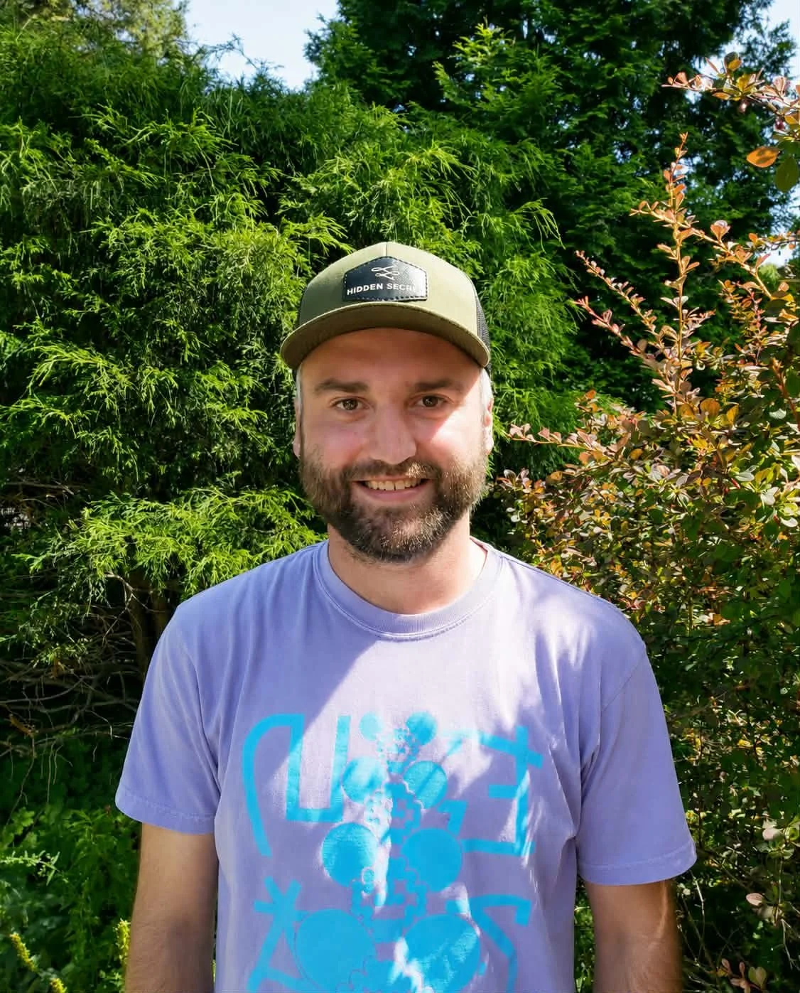 A smiling man with a beard wearing a gray and black baseball cap that says Hidden Secret, and a light purple T-shirt with a blue graphic design, standing outdoors in front of green leafy bushes and trees on a sunny day.