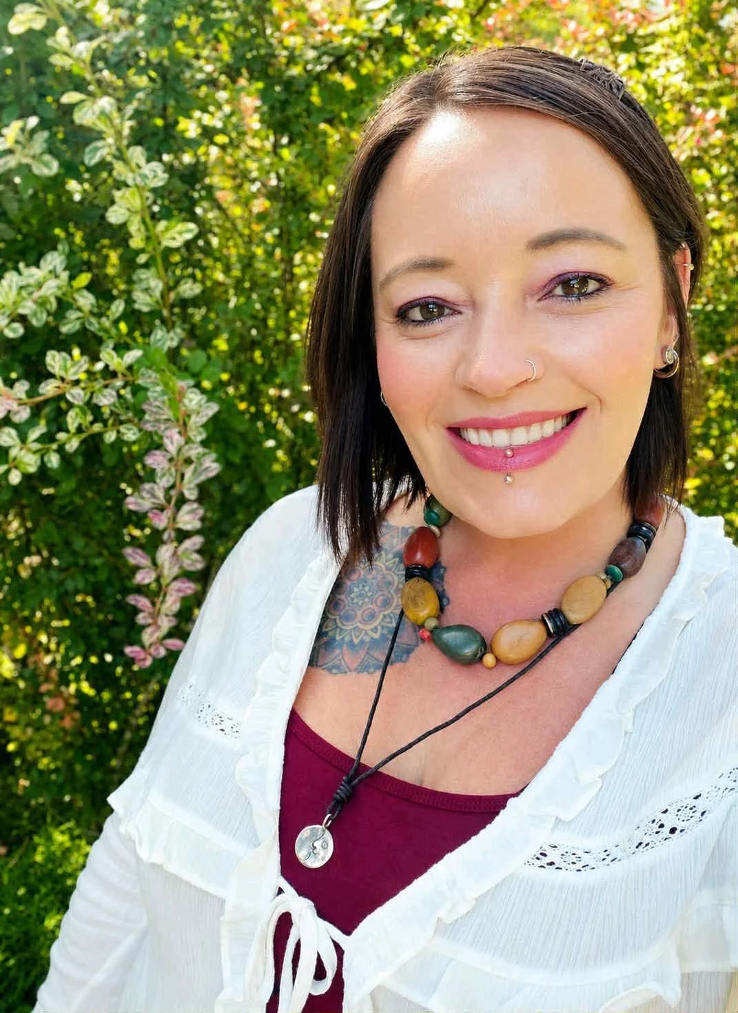 A woman with short dark hair and piercings smiling outdoors, wearing a white blouse and a beaded necklace, with greenery in the background.