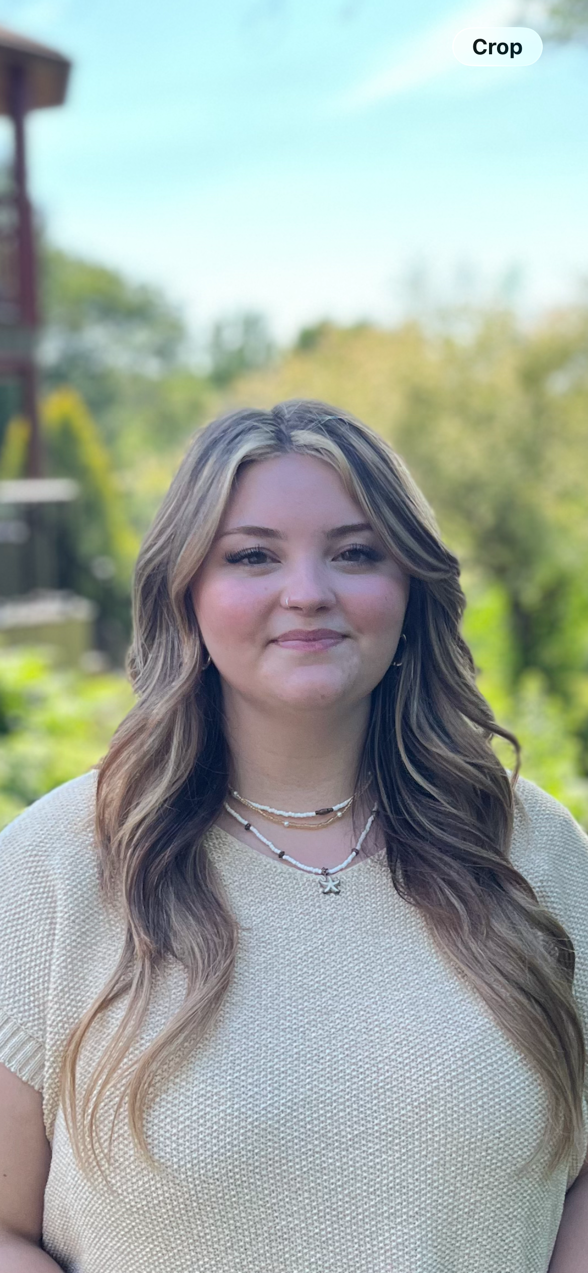 Portrait of a young woman with light brown, wavy hair with blonde highlights, wearing necklaces and a cream-colored textured top, outdoors with green trees and blue sky in the background.