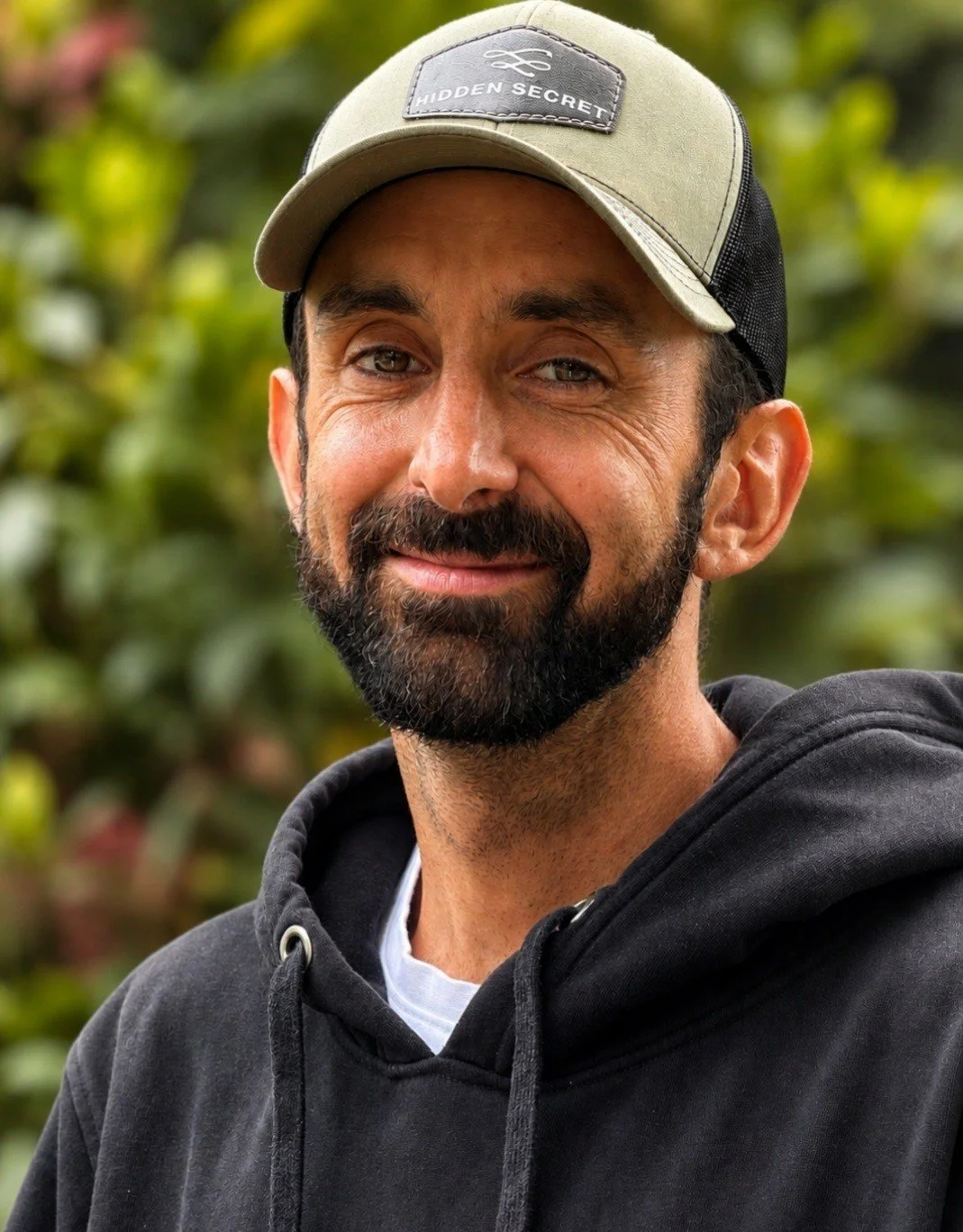 A man with a beard wearing a gray baseball cap and black hoodie, smiling outdoors with green foliage in the background.