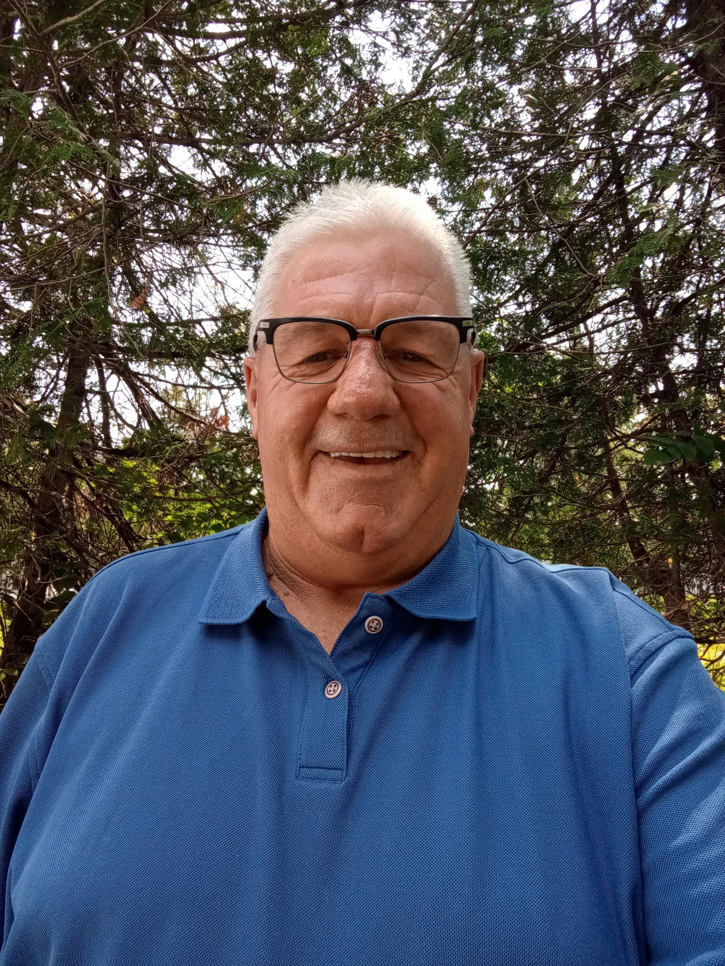 Senior man with gray hair and glasses smiling outdoors, wearing a blue polo shirt with a forested background.